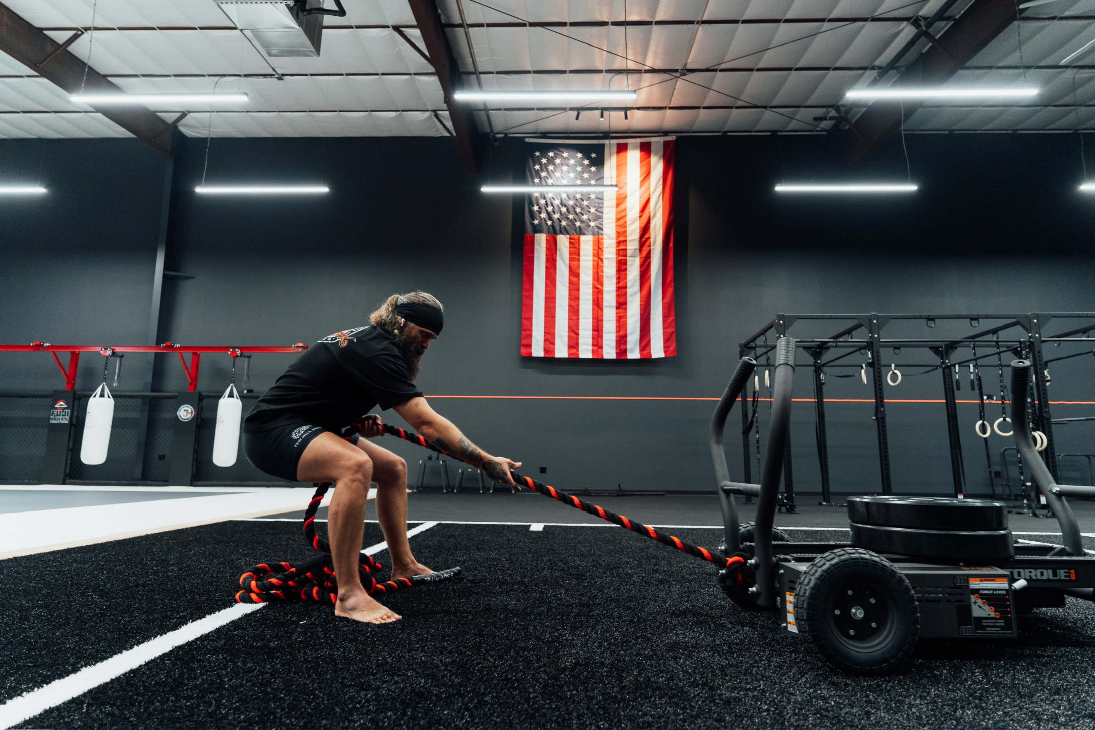 A person pulls a sled with a rope in a gym that features an American flag hanging on the wall.