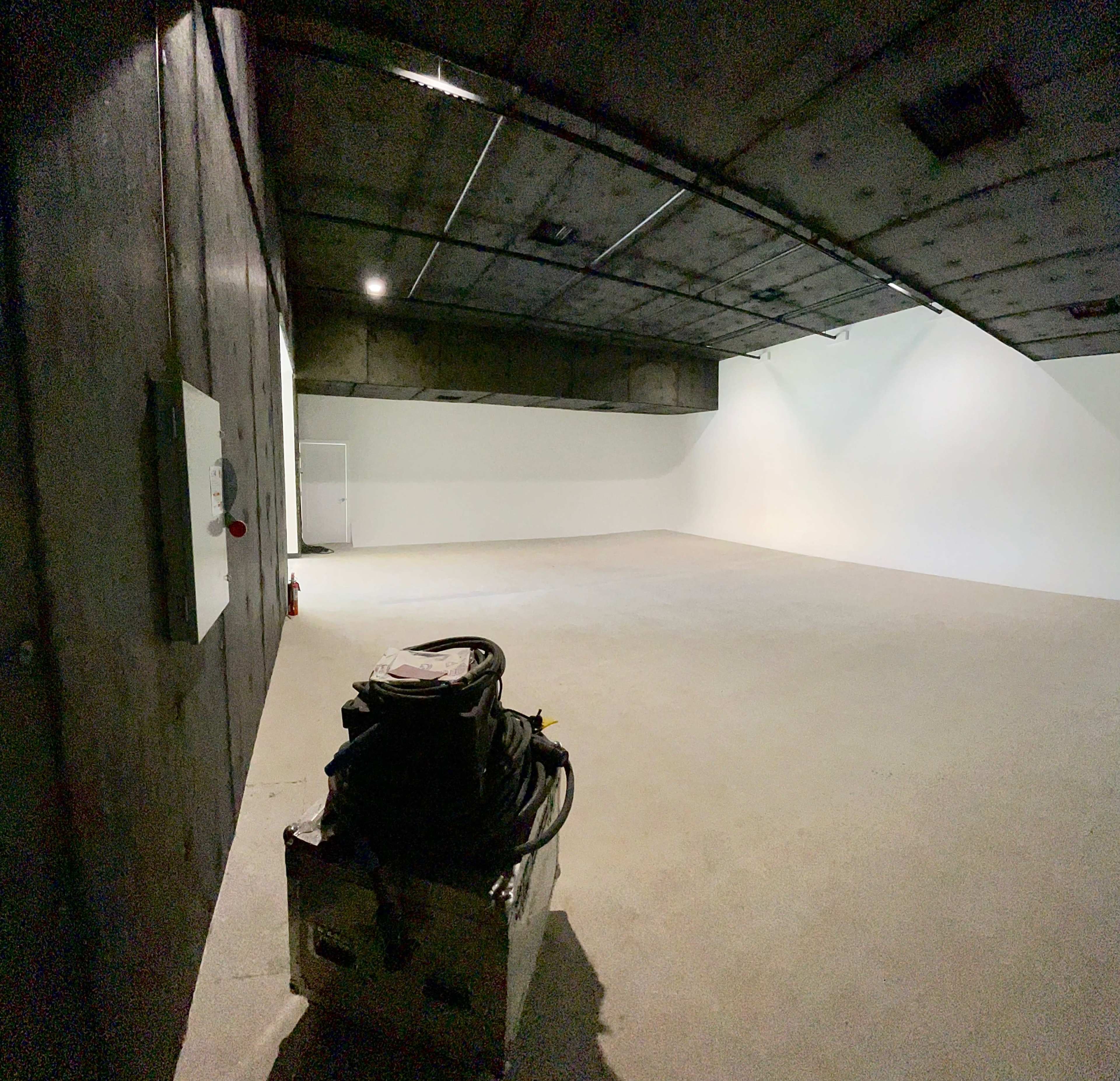 An empty, dimly lit room with concrete walls and a smooth floor, featuring a bag and equipment in the foreground.