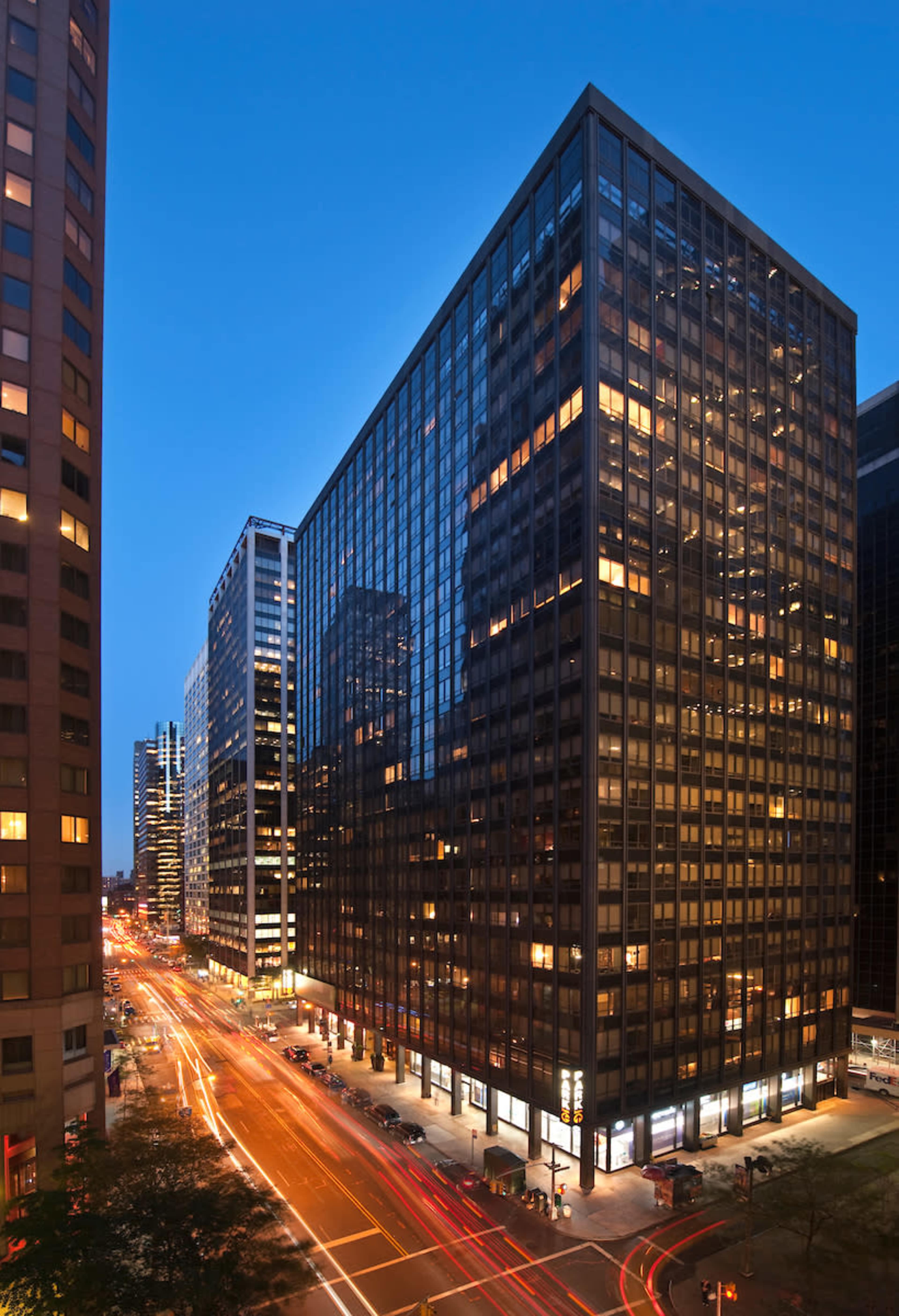 The image shows a city street with tall glass skyscrapers on either side, illuminated by the evening lights and streaks of car headlights.