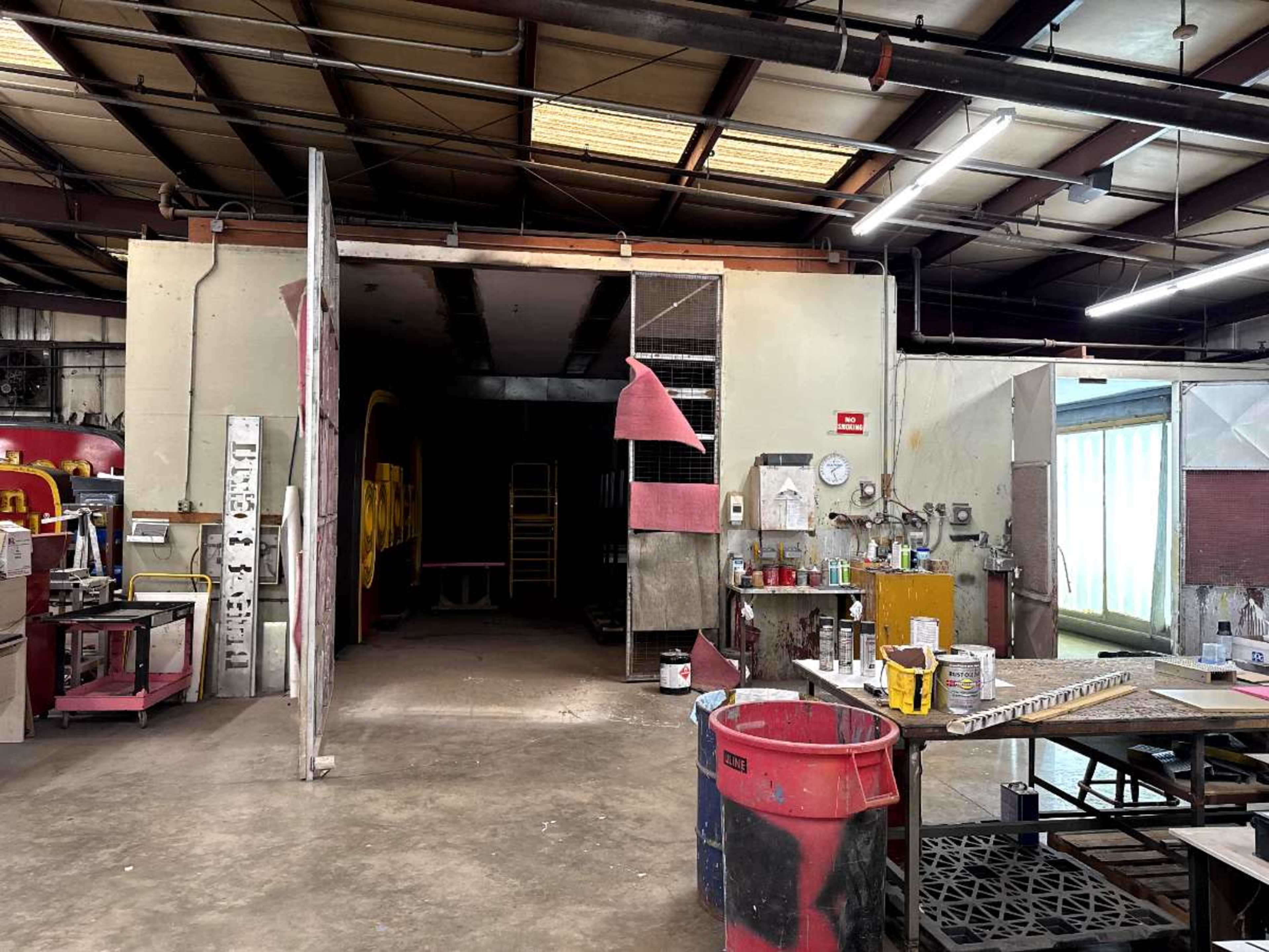 The image shows a spacious workshop with various tools, equipment, and paint supplies organized along the walls, and an open doorway leading to a darker area.
