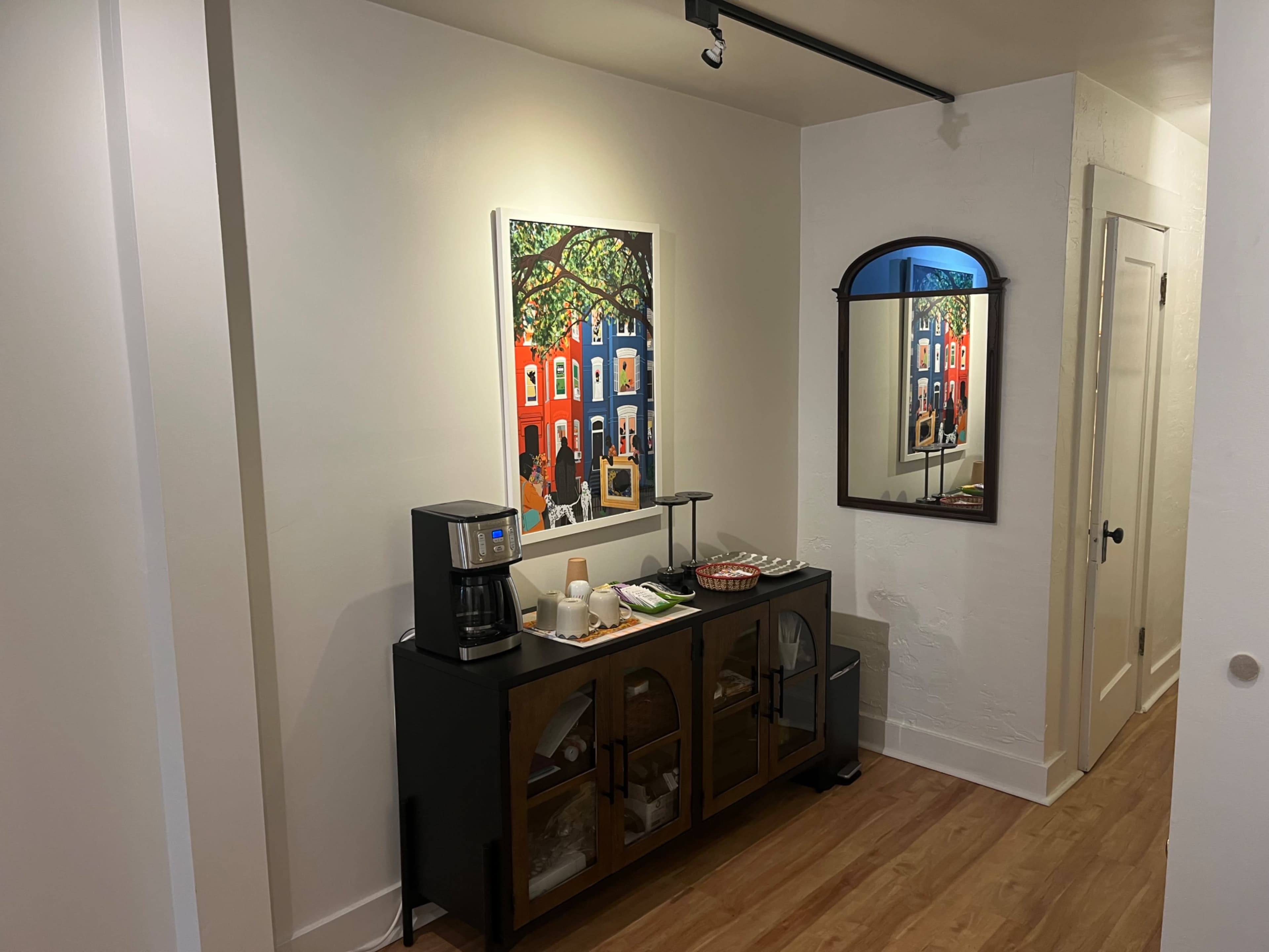 The image depicts a well-lit hallway featuring a colorful painting on the wall, a wooden cabinet with glass doors, a coffee maker, and a mirror reflecting the surroundings.