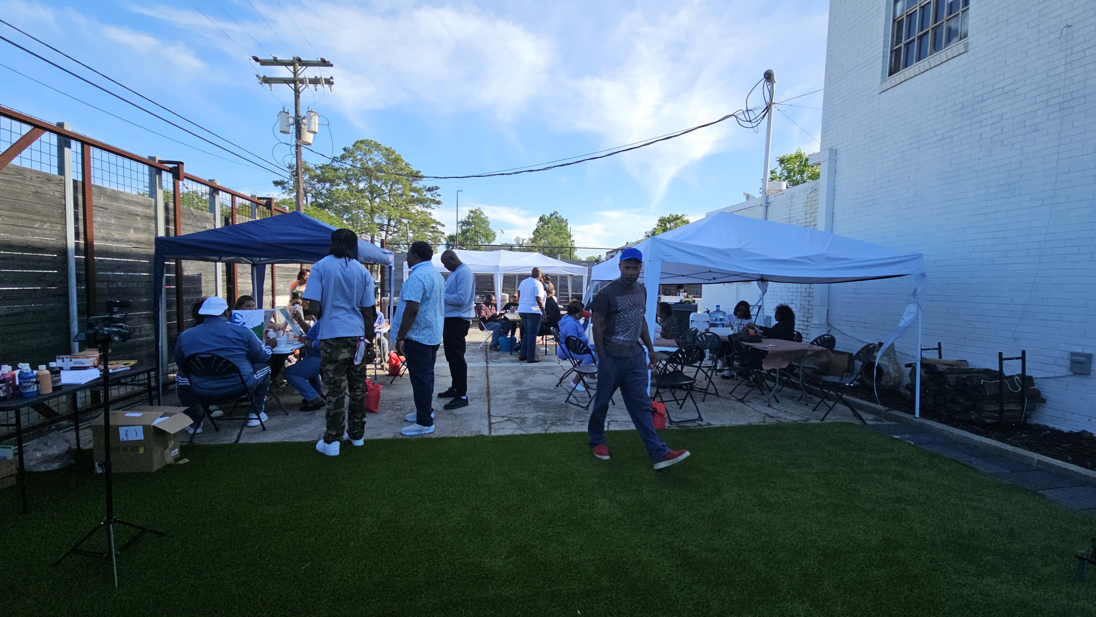 The image shows a gathering taking place outdoors, featuring several tents, people socializing, and chairs arranged on a grassy area.