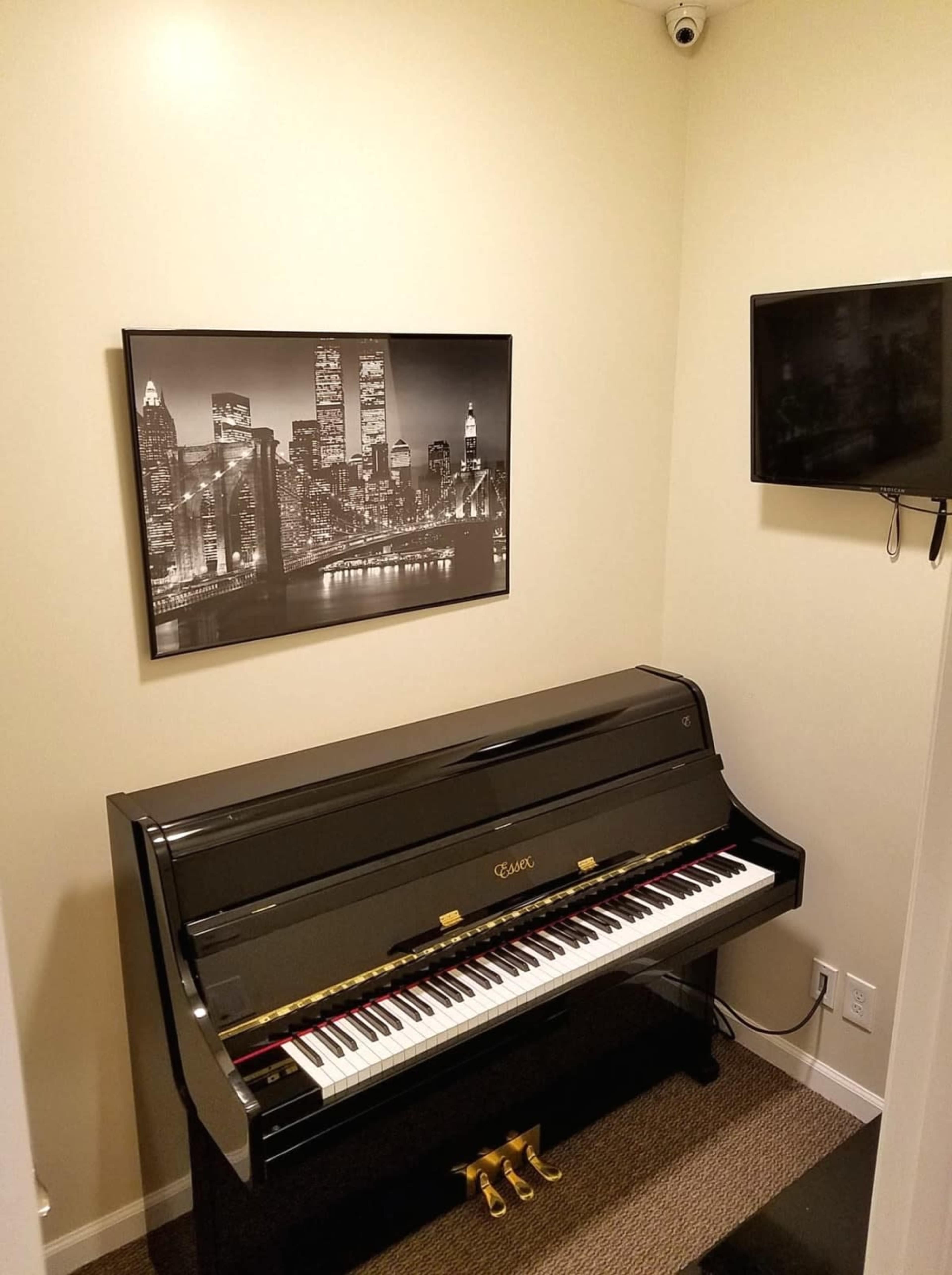 A black piano is positioned against a wall in a small room, featuring a black-and-white cityscape photograph and a mounted television nearby.
