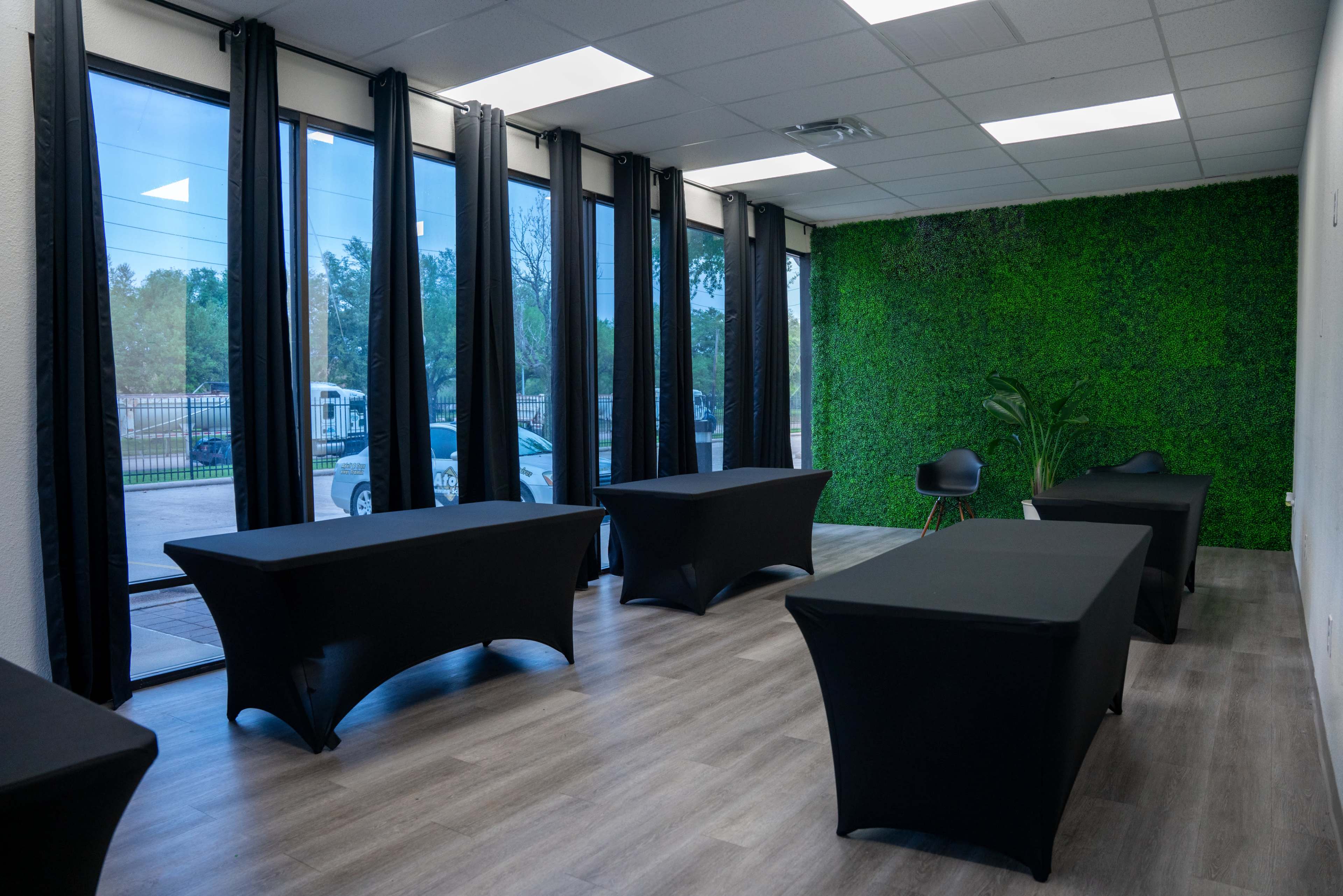The room features several black tablecloth-covered tables arranged in a bright space with large windows and a green plant wall.