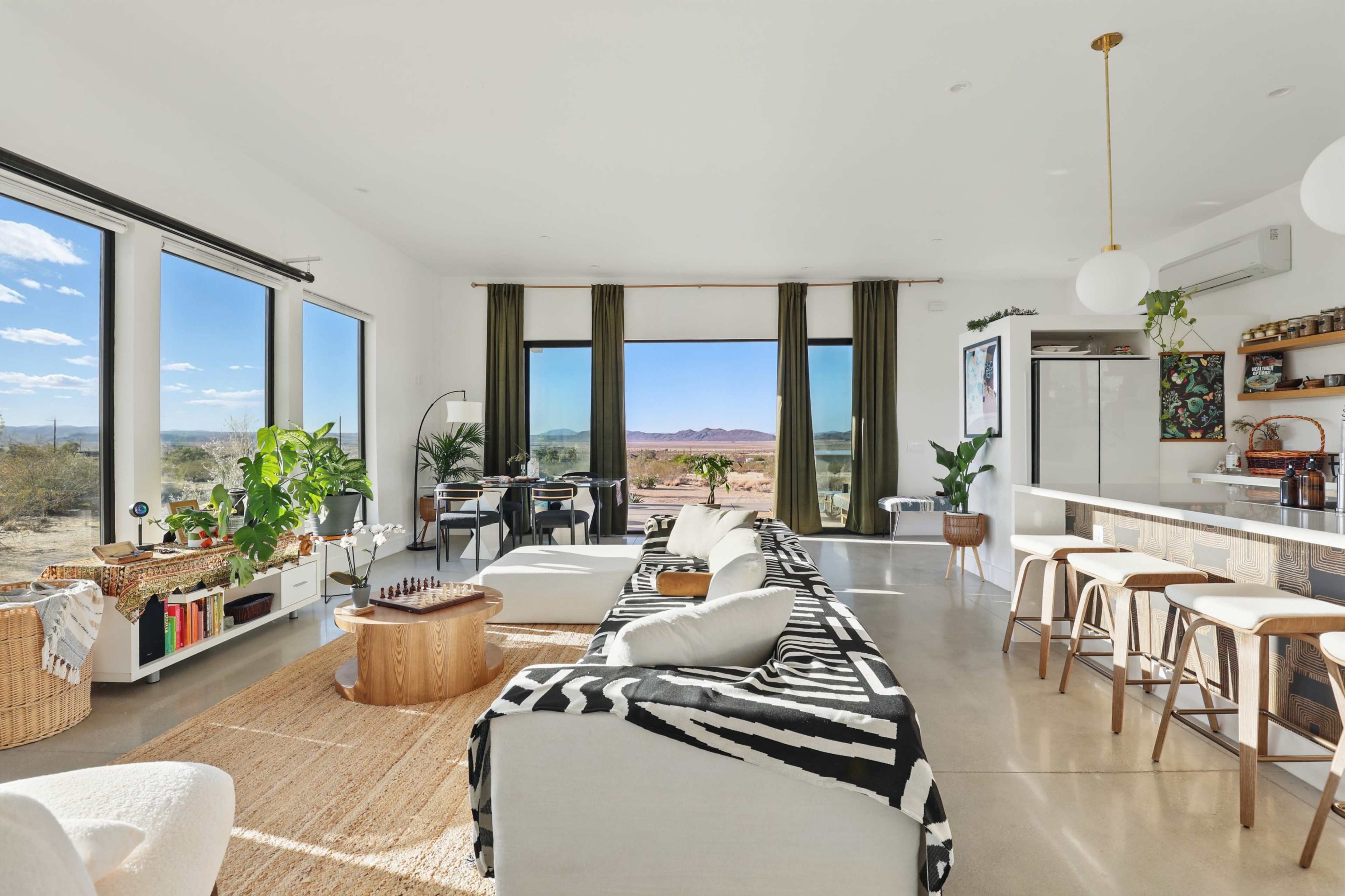 The spacious living area features large windows with a view of the desert landscape, a modern kitchen island, and a seating arrangement with a patterned sofa and a round rug.