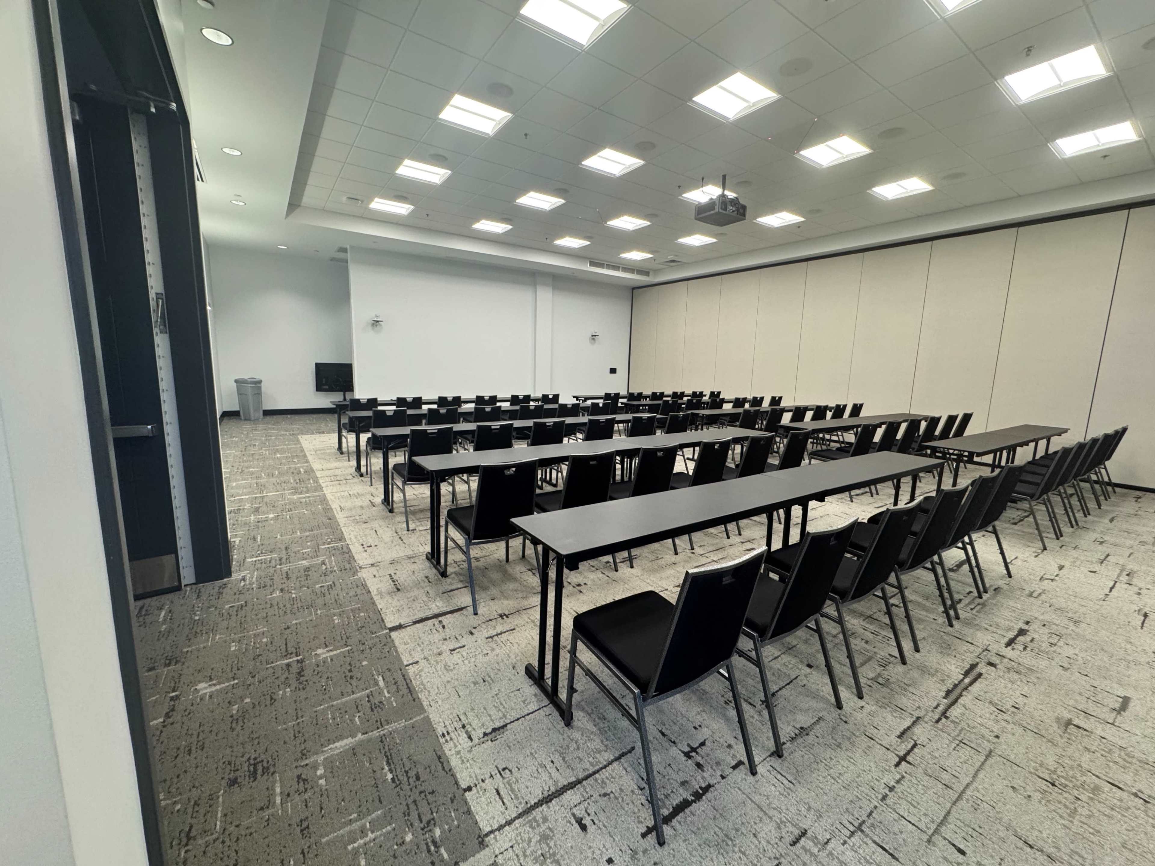 A conference room features rows of black tables and chairs arranged in a grid pattern, with light-colored walls and a projector mounted on the ceiling.