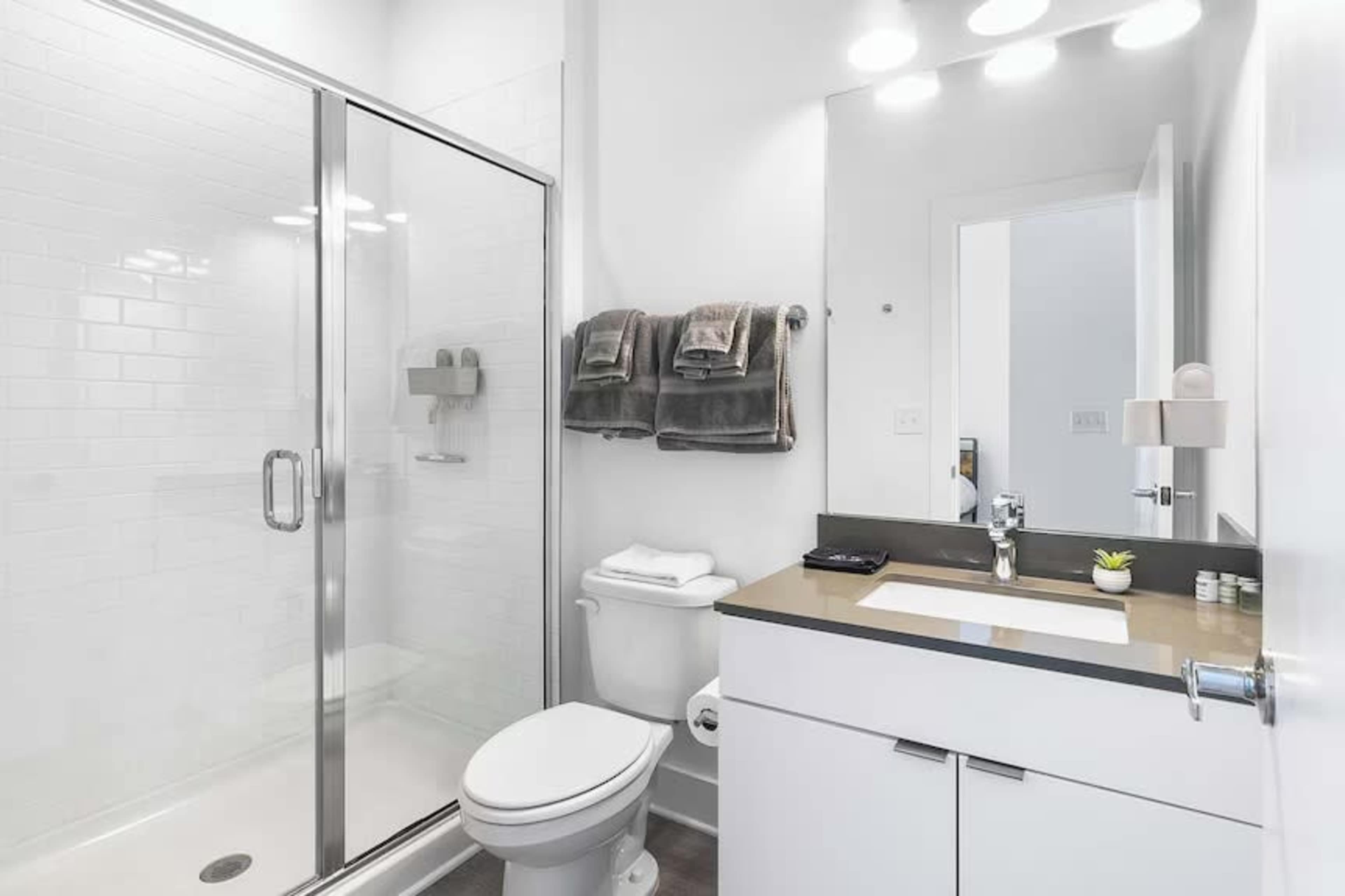 The image shows a modern bathroom with a glass shower, a toilet, a sink with a countertop, and neatly arranged towels.