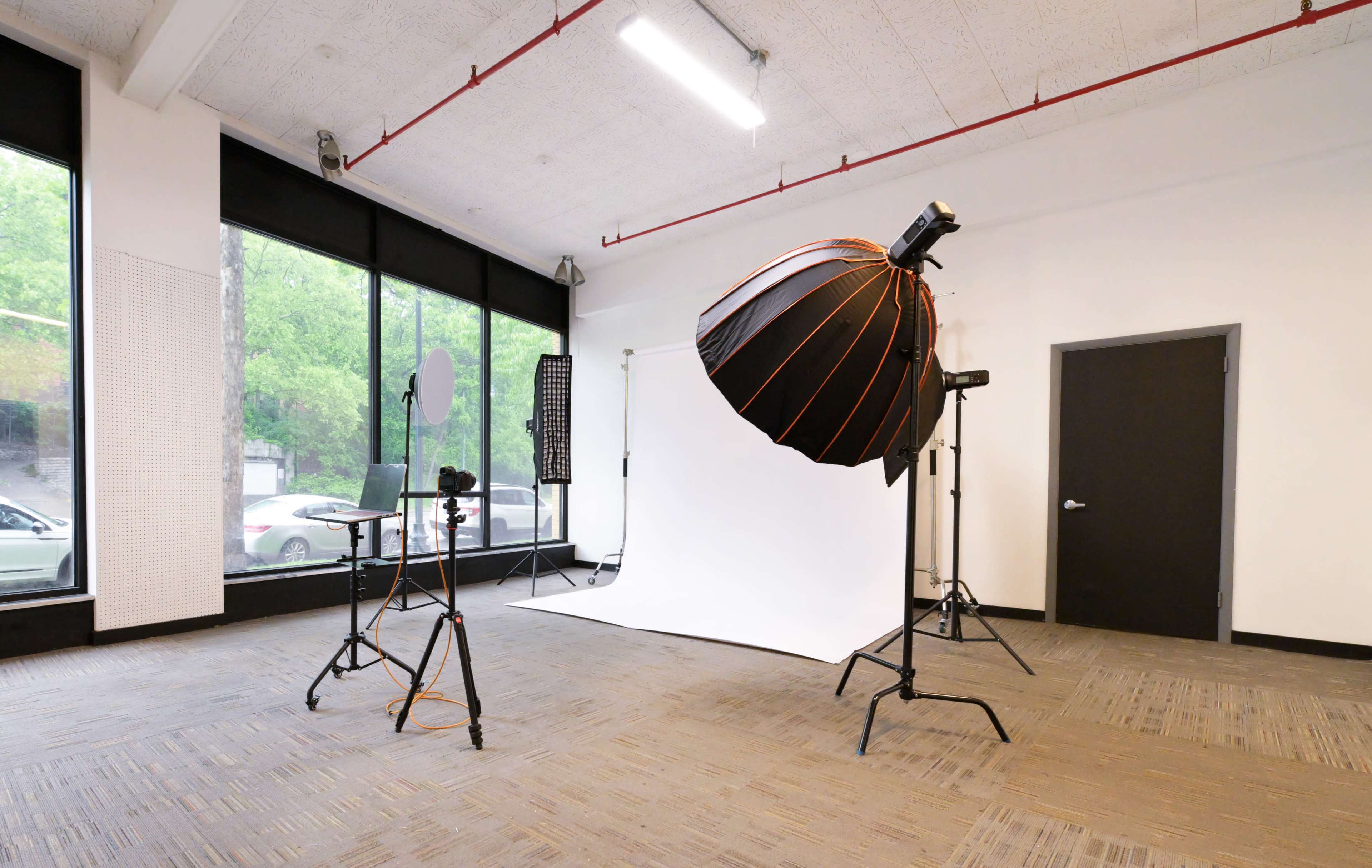 The image shows a photography studio with a large softbox light, a white backdrop, and equipment set up on tripods.