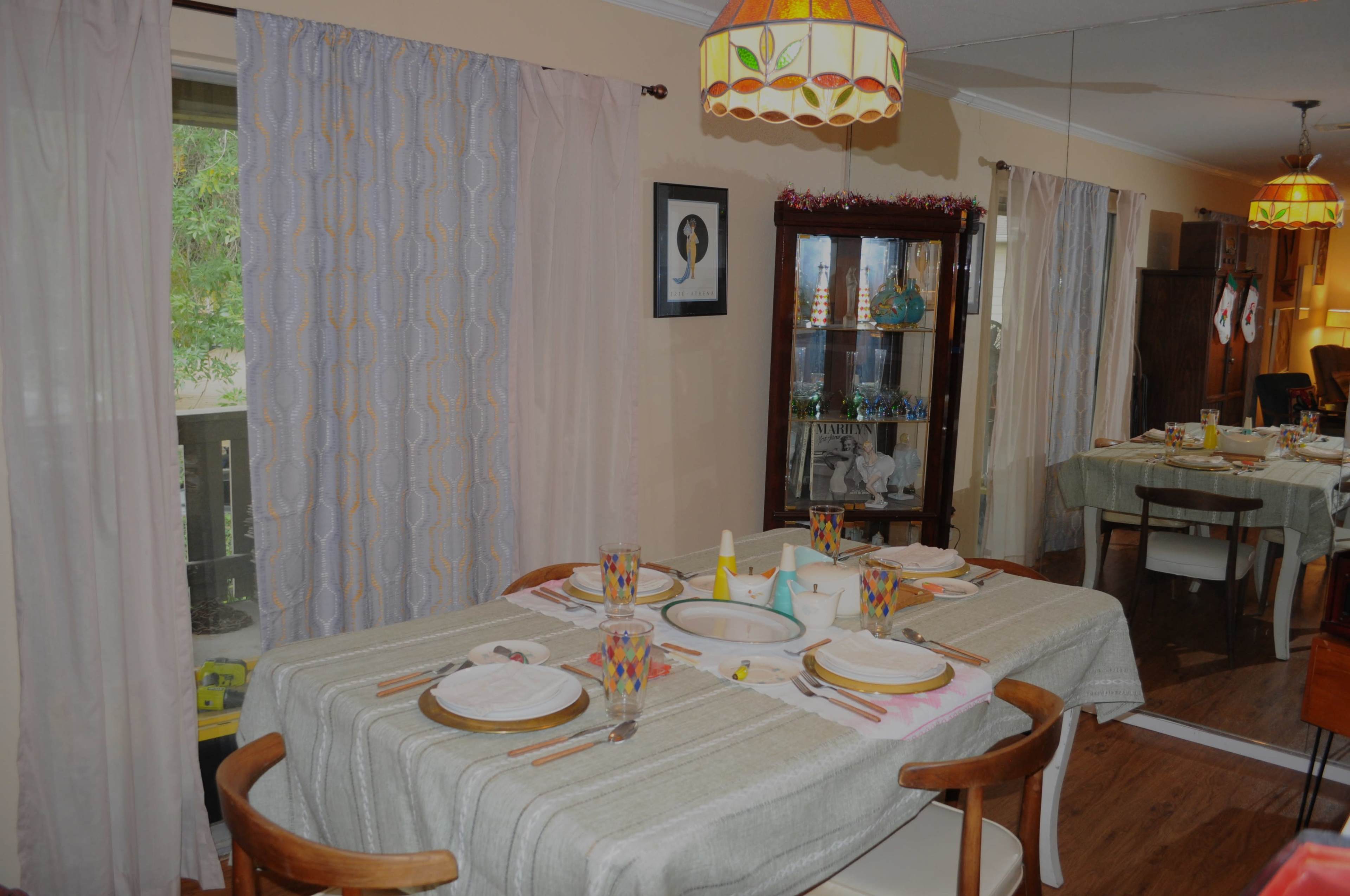 The image shows a dining room with a table set for a meal, featuring plates, glasses, and a centrally placed bowl, surrounded by chairs and a cabinet displaying decorative items.