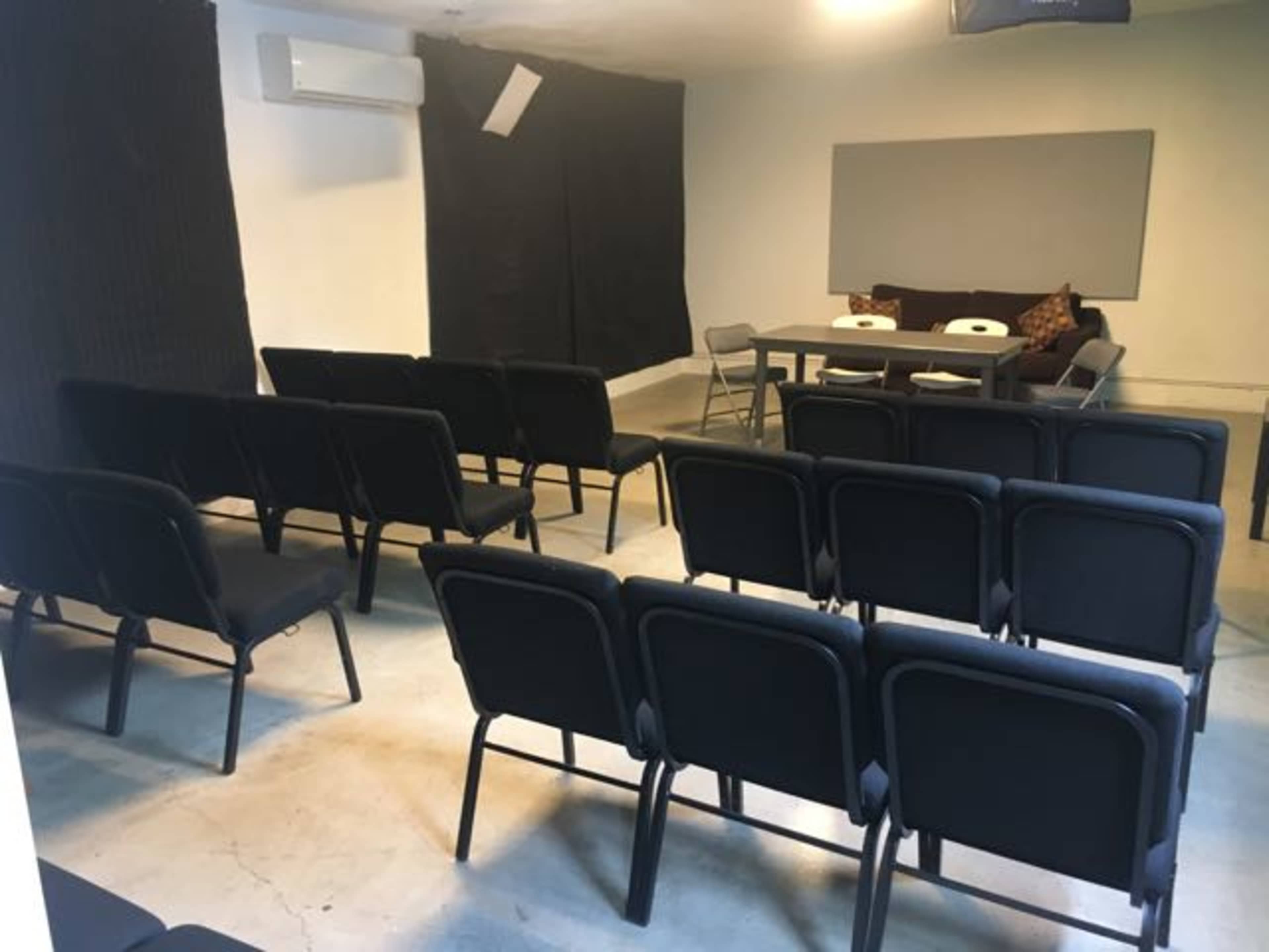 A room with a row of black chairs facing a table and a wall, with additional chairs arranged to the side.