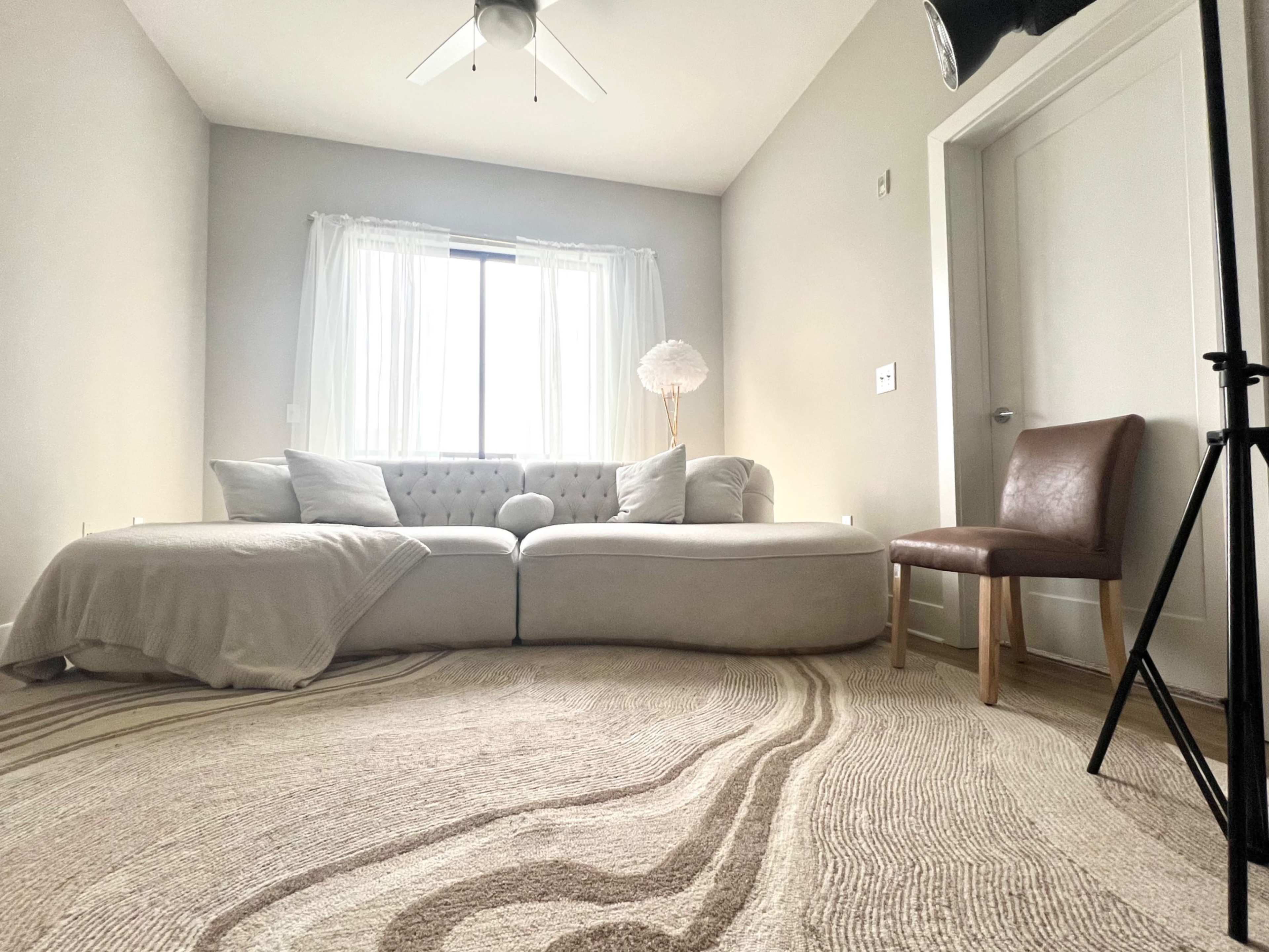 A modern living room features a curved white sofa, a brown chair, and a lamp, with large windows allowing natural light to enter.