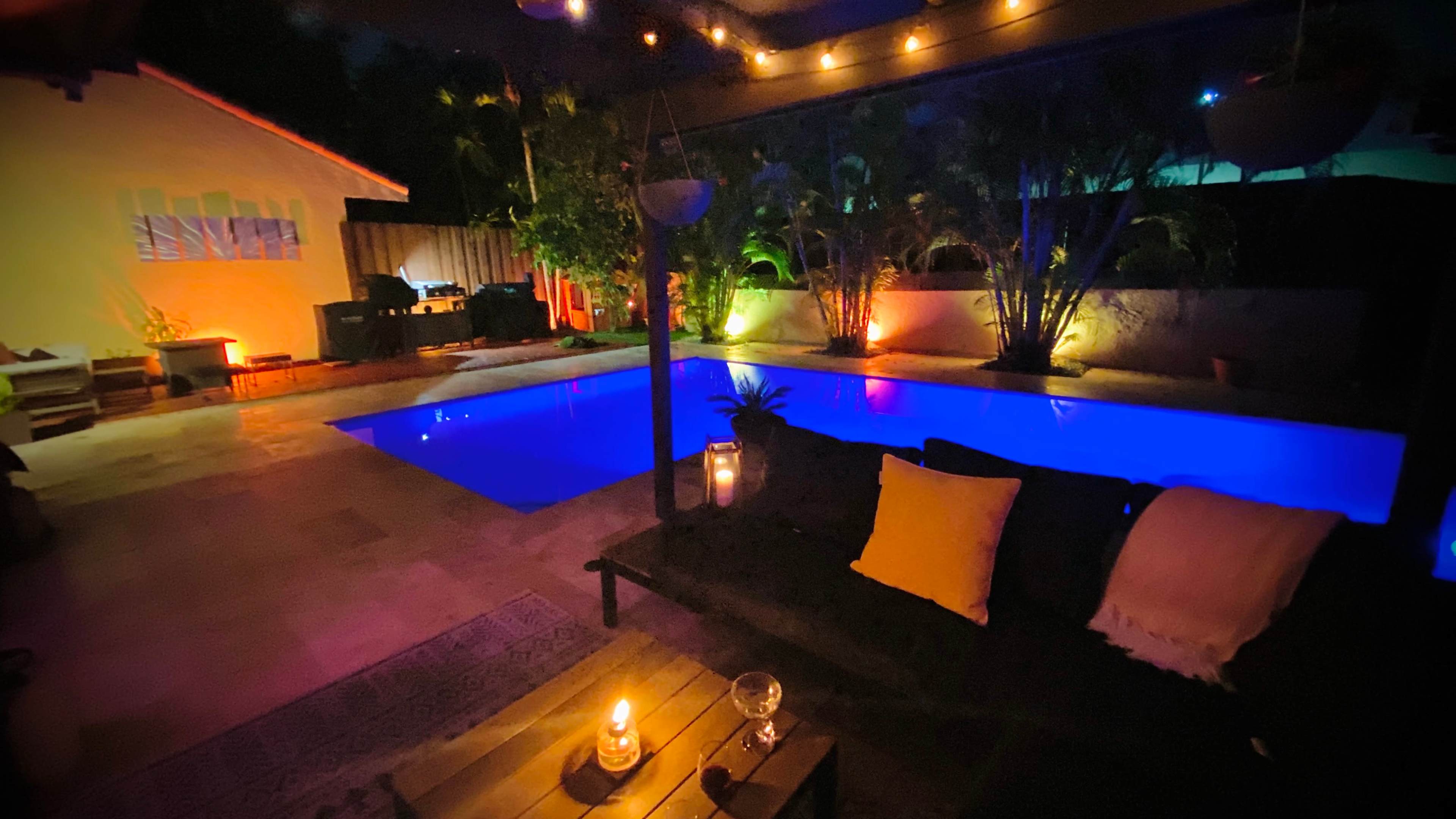 The image shows a well-lit outdoor pool area at night with lounge seating and decorative plants.