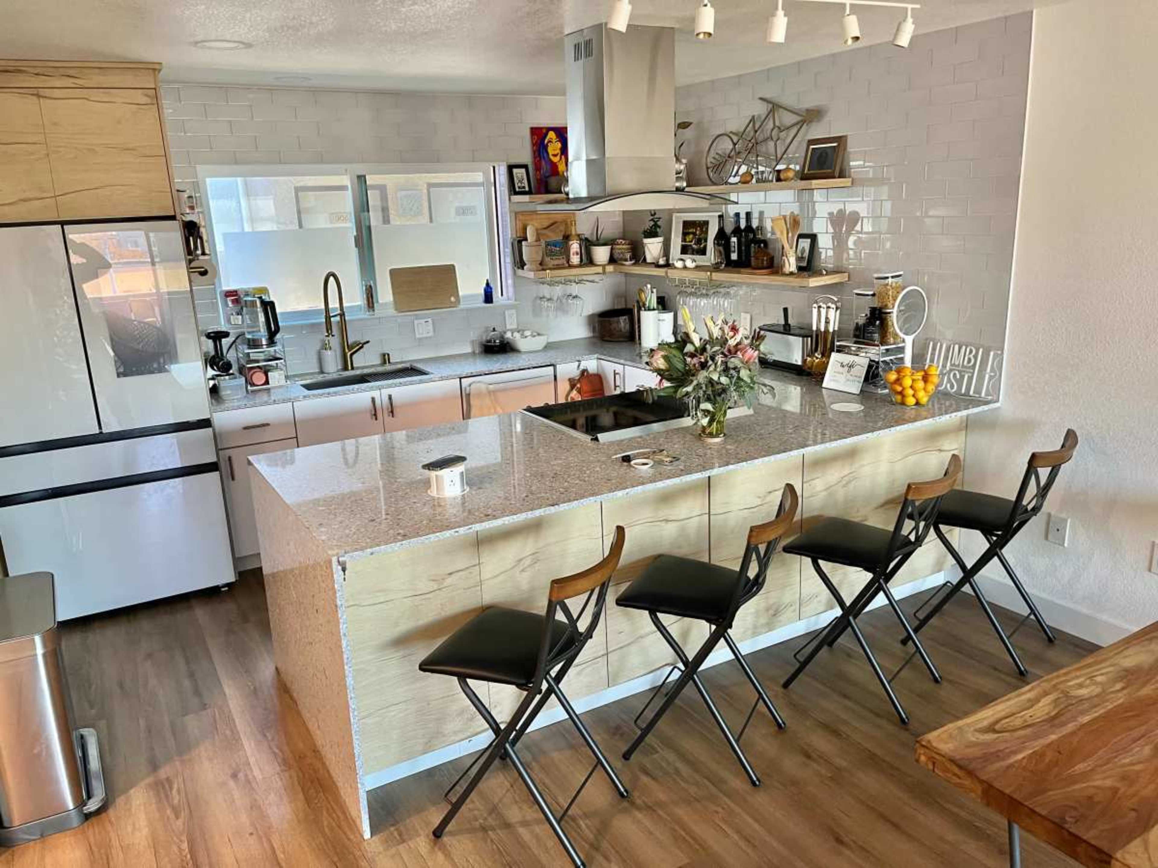A modern kitchen with a center island, four black stools, and various kitchen appliances and decor on the shelves.