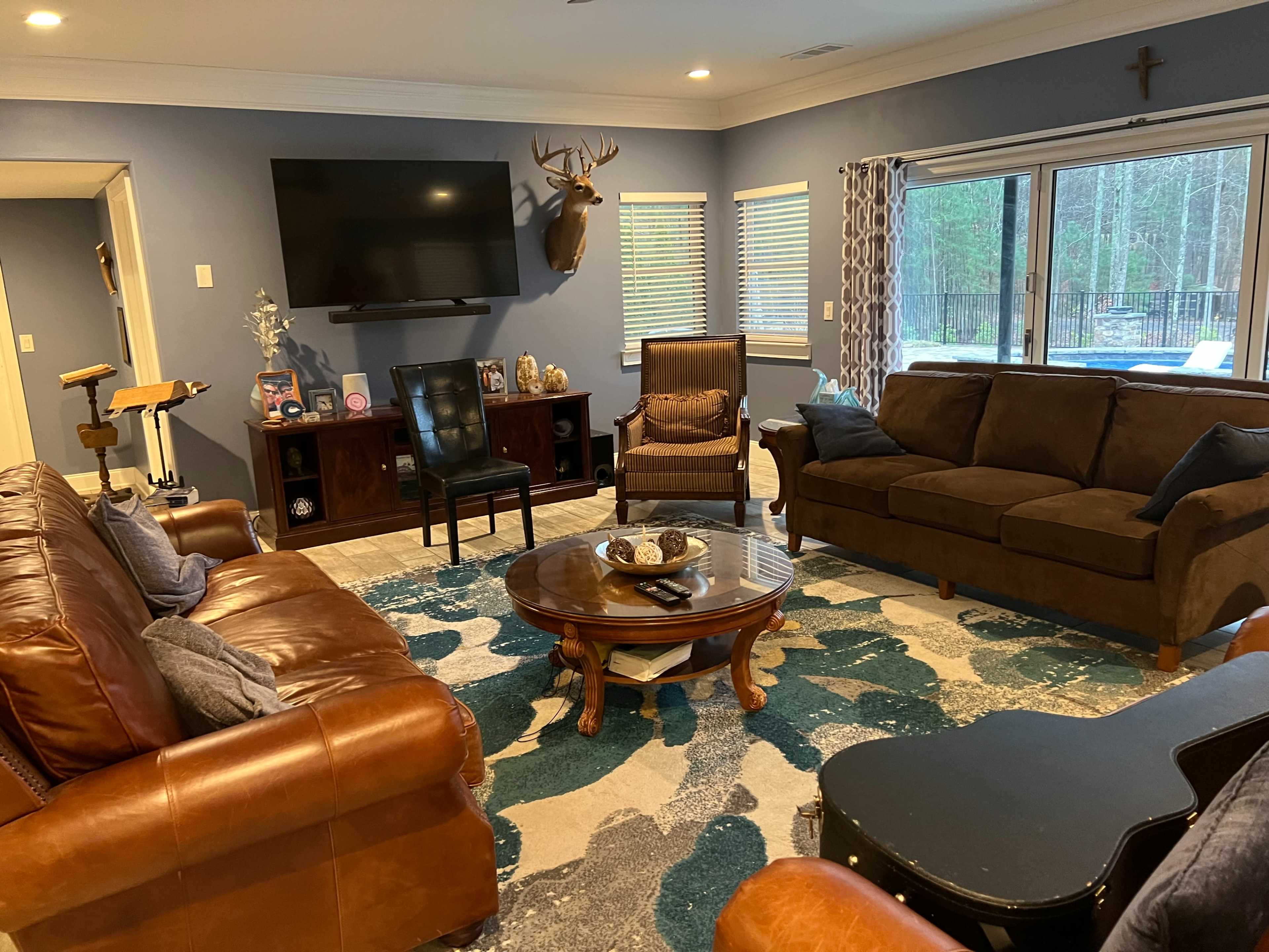 A living room with a large television mounted on the wall, a mounted deer head, and a mix of leather and fabric furniture arranged around a coffee table on a patterned rug.