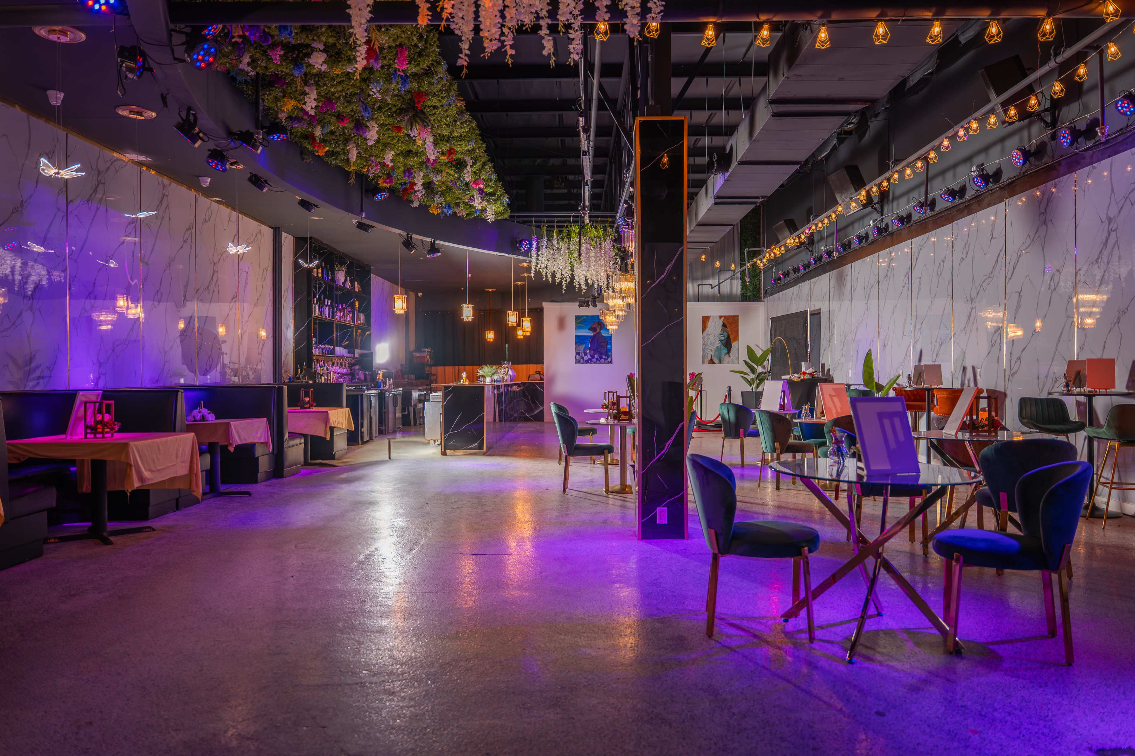 The image shows a modern restaurant interior with dim lighting, featuring tables and chairs arranged in a spacious layout, along with decorative plants and hanging lights.