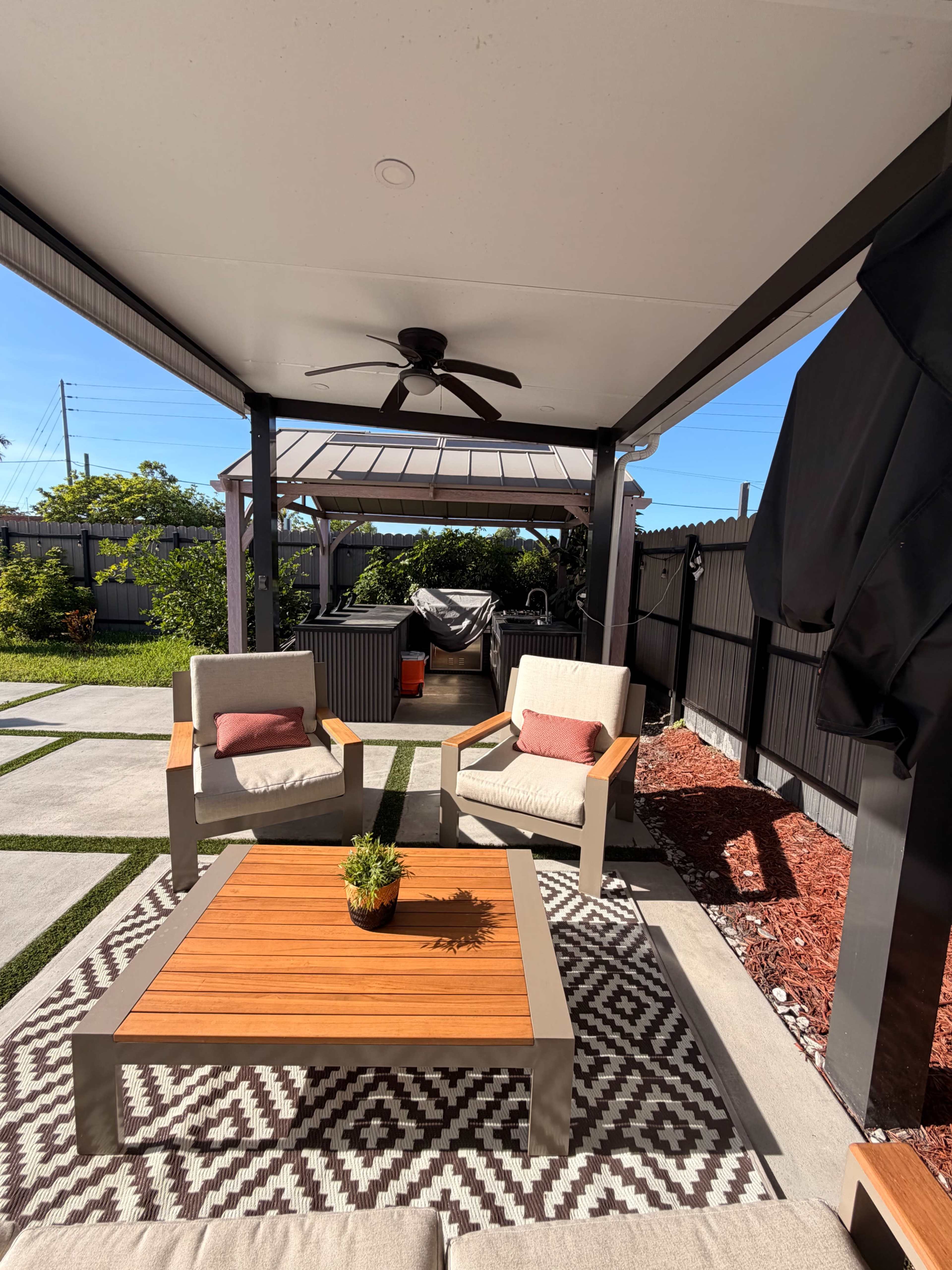 A covered outdoor seating area features two chairs and a wooden table, with a gazebo and grill visible in the background.