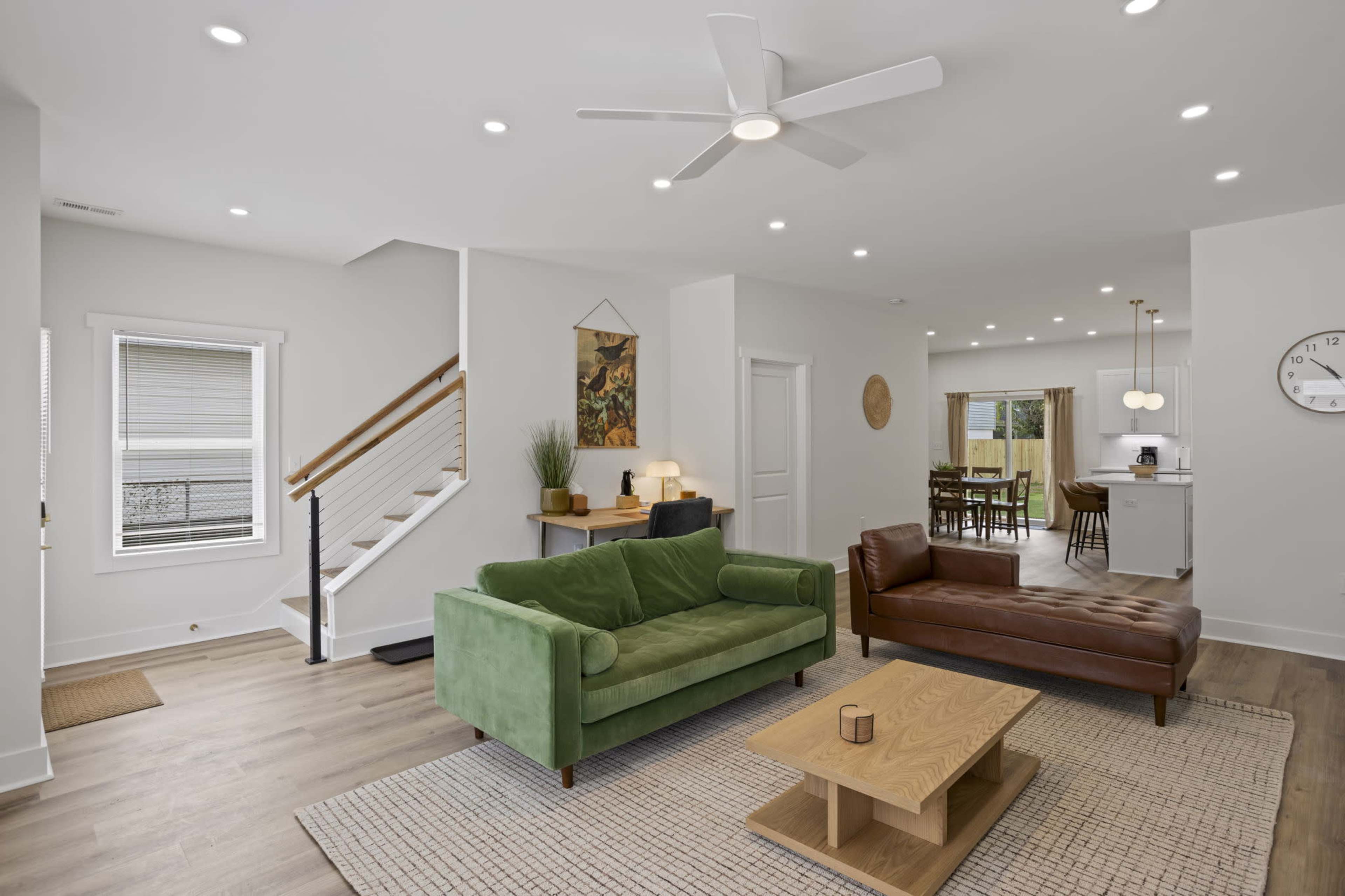 The image shows a modern living room featuring a green sofa, a brown leather chair, a wooden coffee table, and a staircase leading to an upper level.