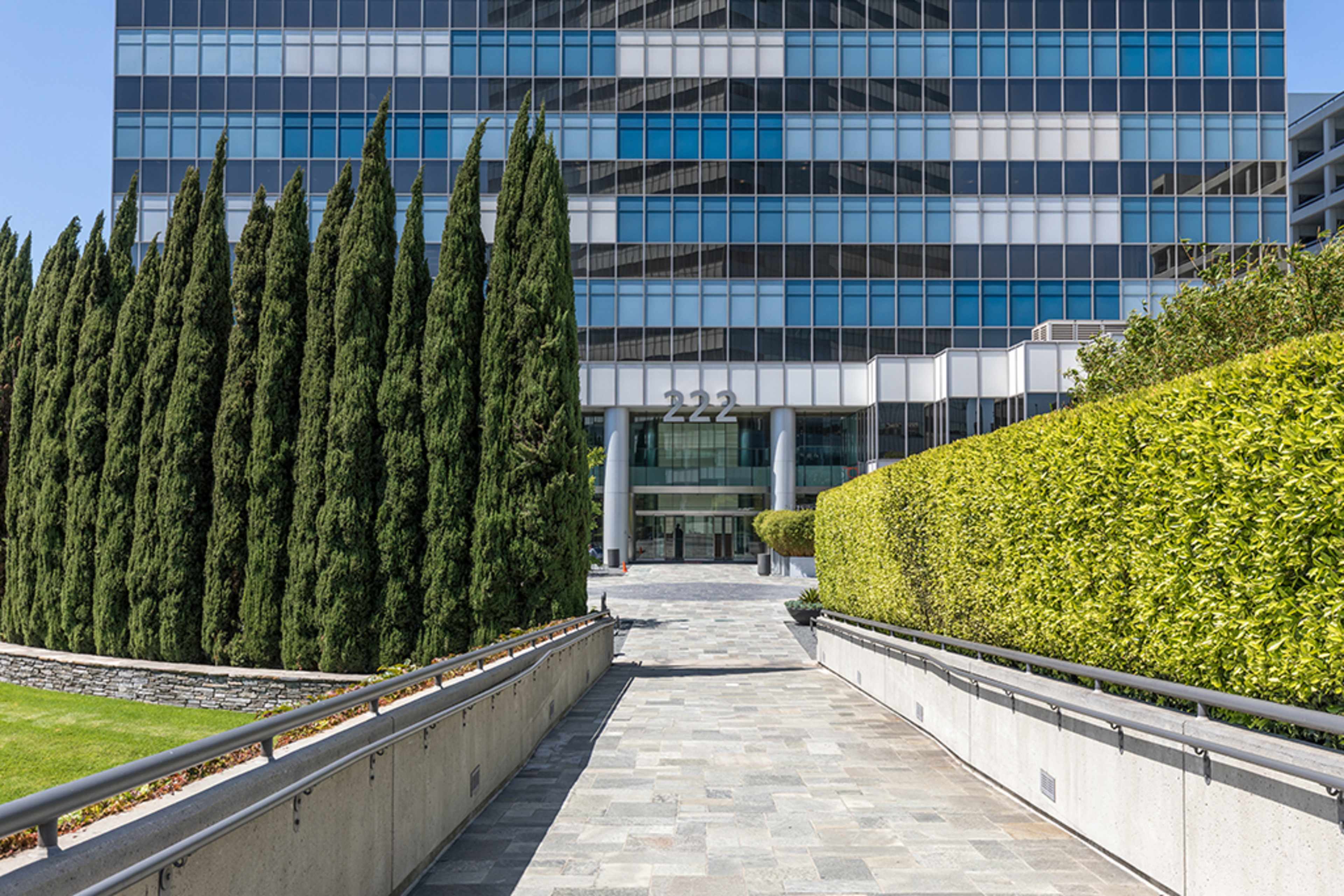 A stone pathway leads to a modern glass office building with the number "222" prominently displayed at the entrance, flanked by tall cypress trees and manicured hedges.