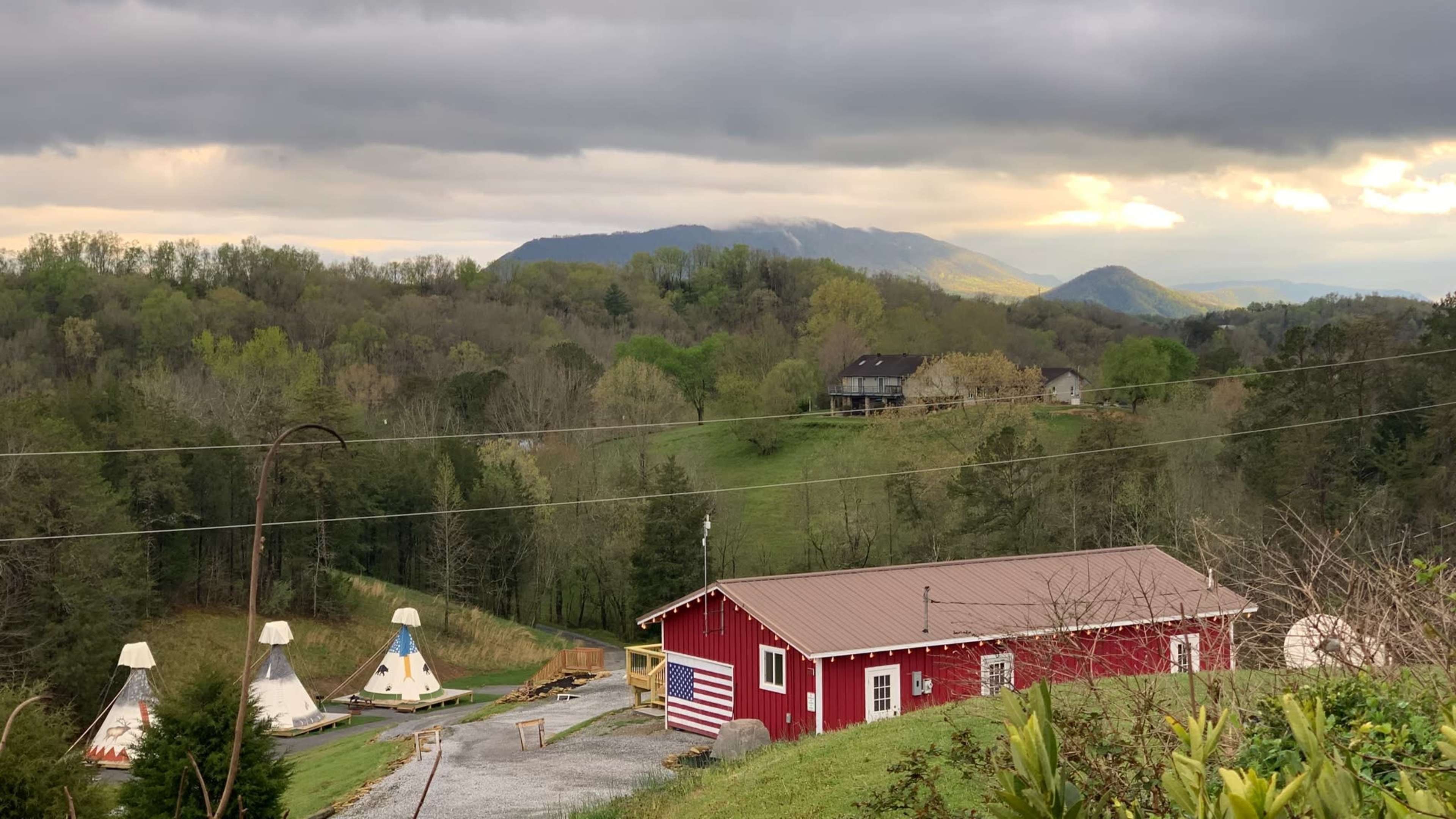 A red barn with an American flag stands near three teepees on a hilly landscape under a cloudy sky.