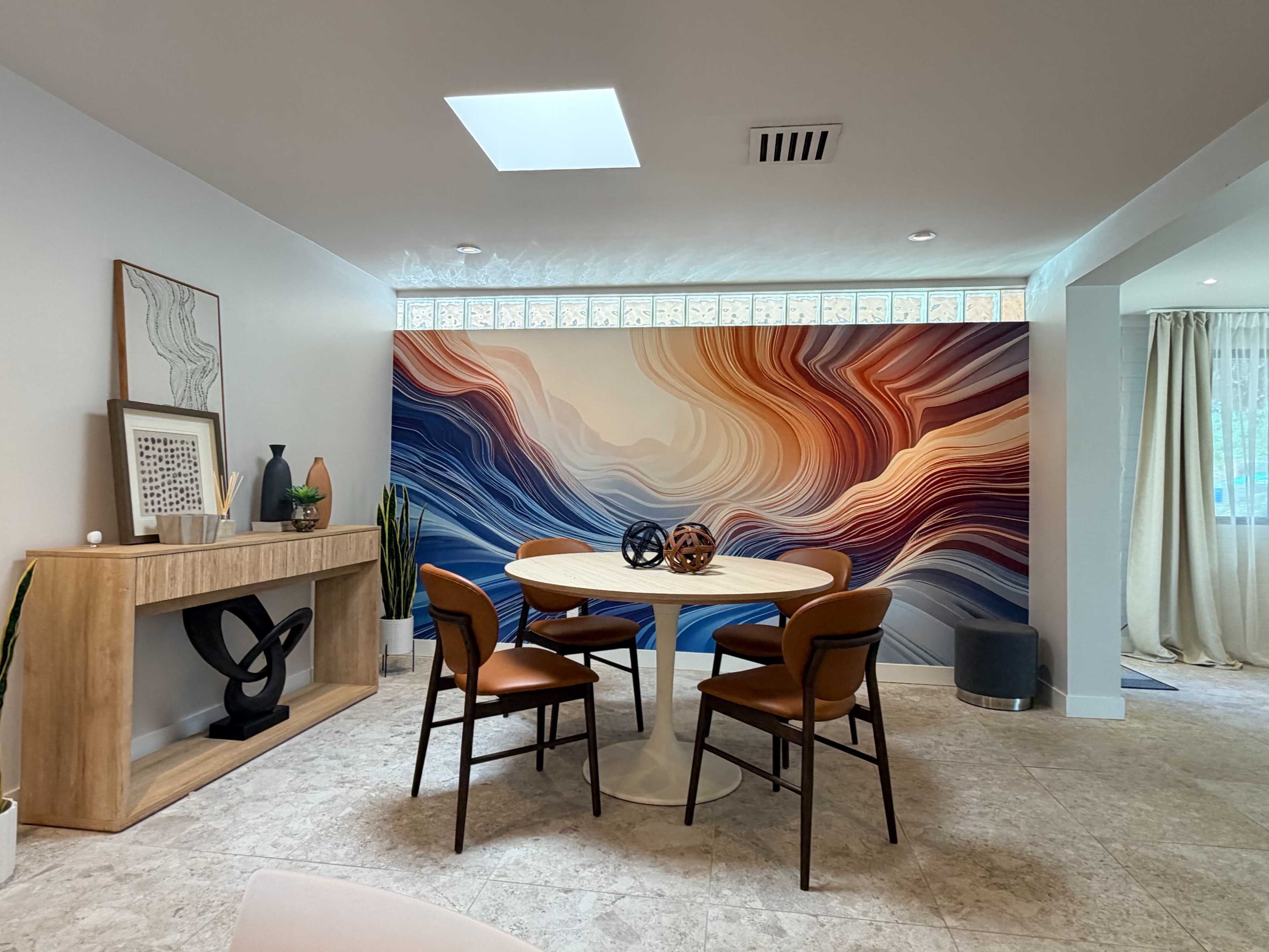 A dining area features a round table surrounded by four chairs, with a colorful mural on the wall and a wooden sideboard.