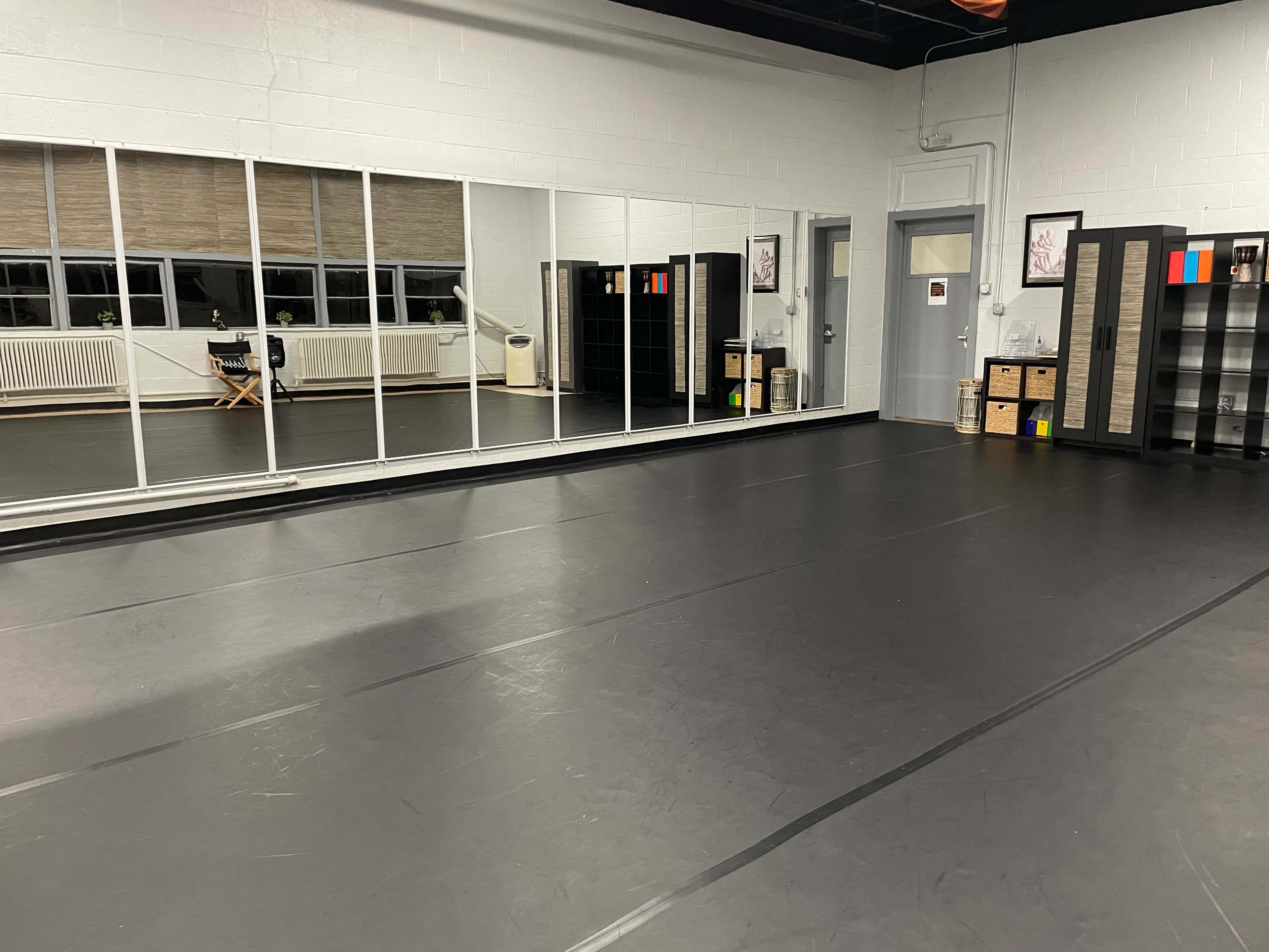 A spacious dance studio with mirrored walls, a black floor, and storage furniture along one side.