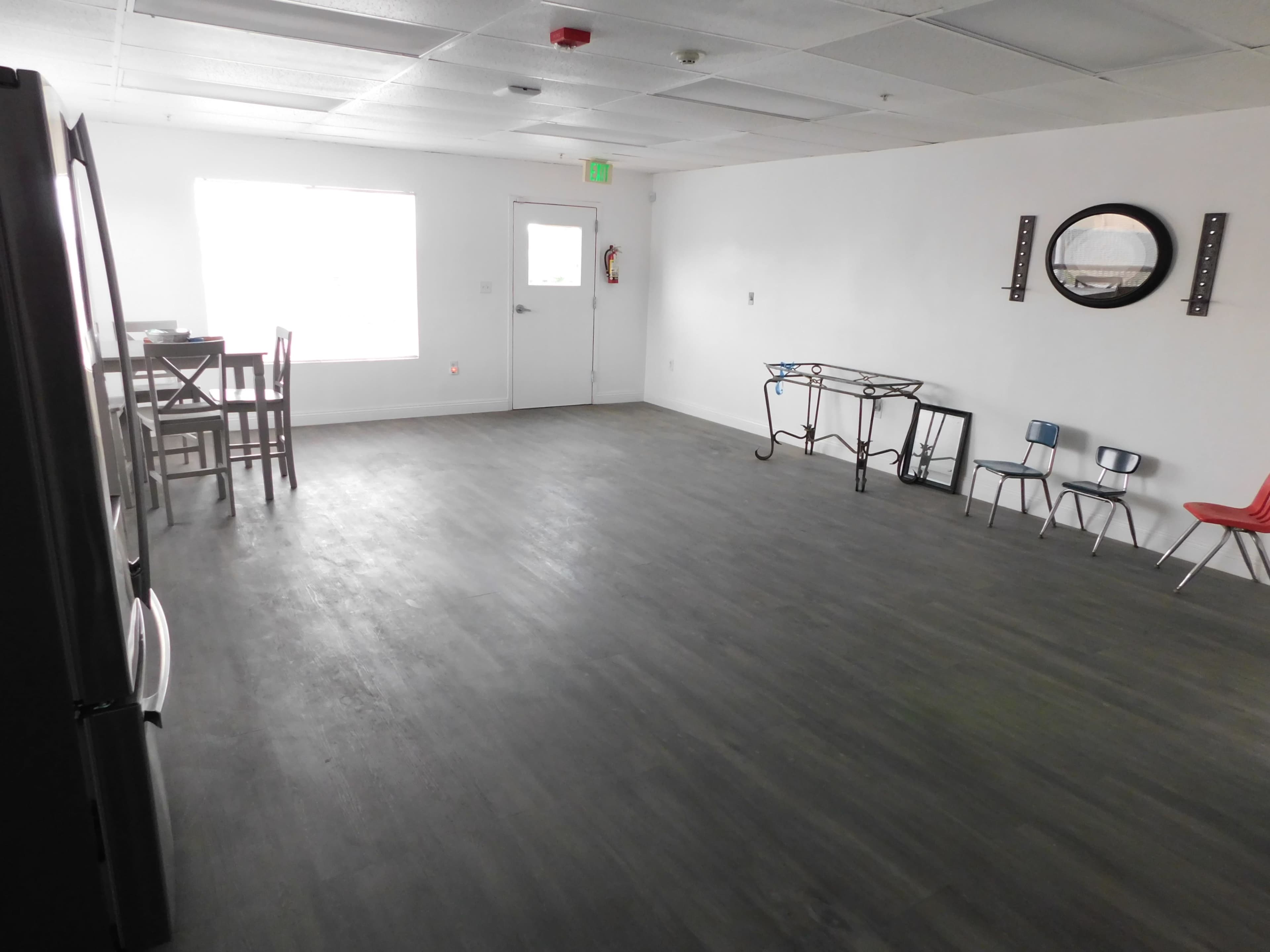 The image shows a large, empty room with gray flooring, a window, and minimal furniture including a table and chairs.