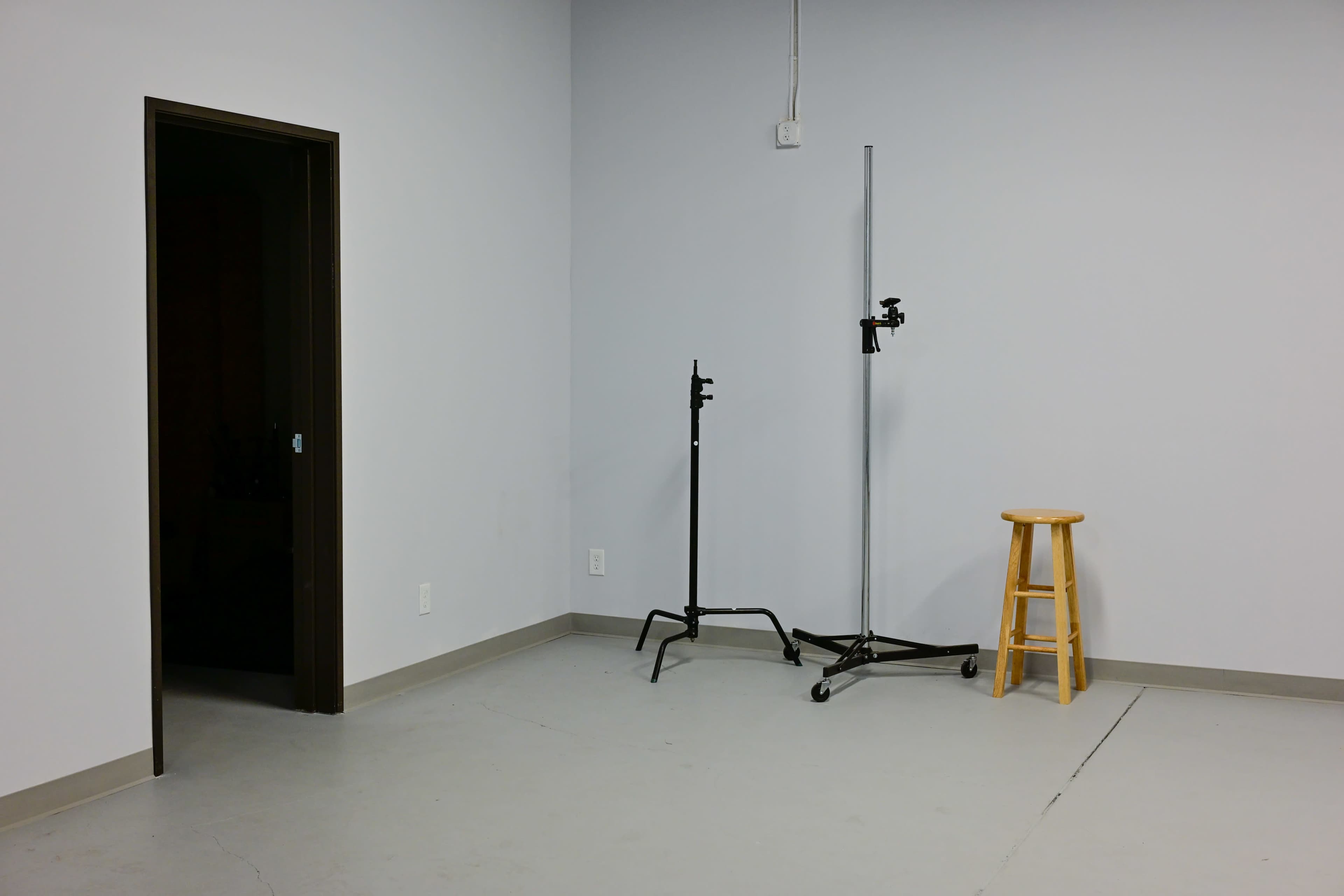 The image shows a sparse room with a gray wall, a door on the left, and two stands next to a wooden stool on the right.