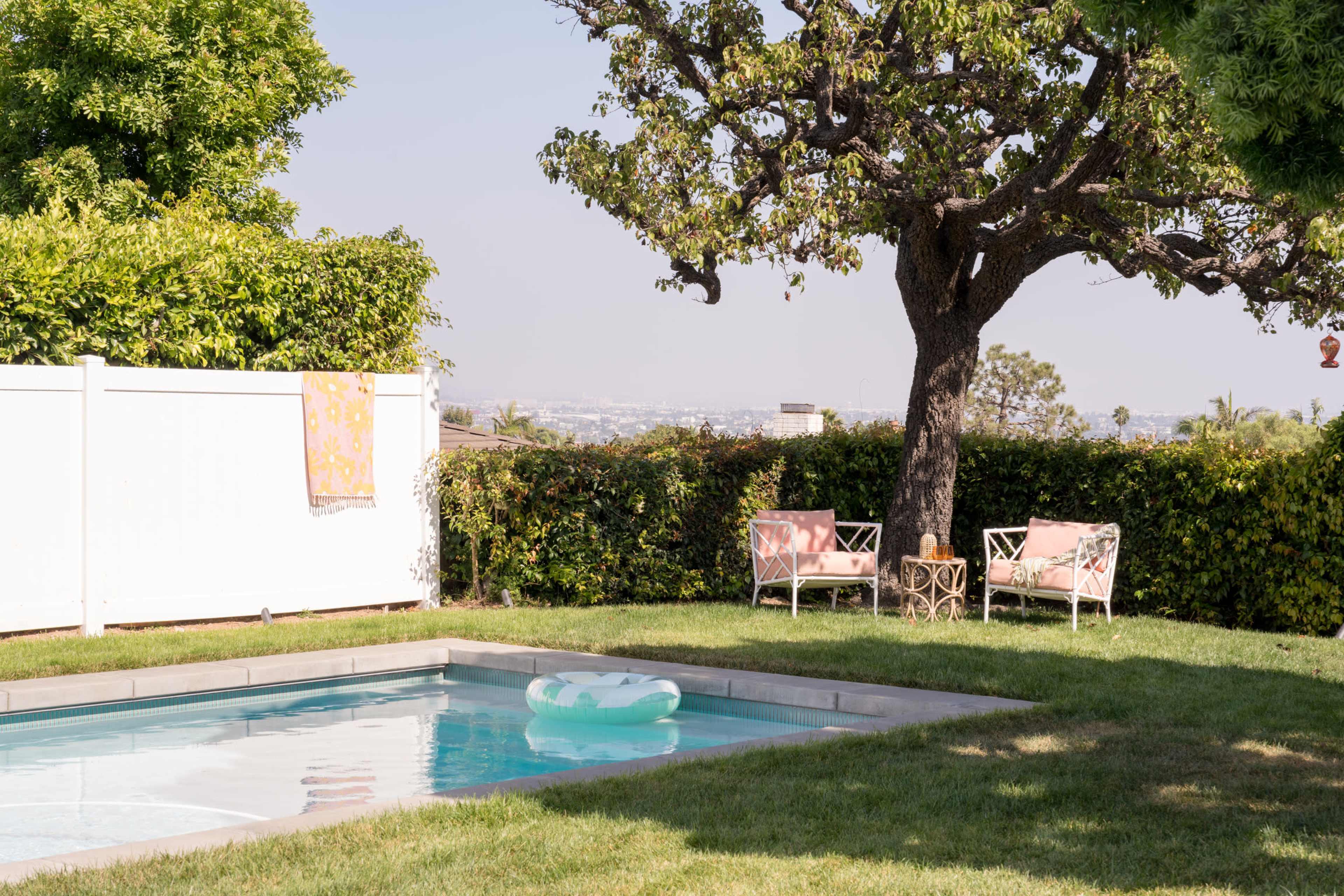 A clear swimming pool is surrounded by a grassy area and a large tree, with two chairs placed nearby and a colorful towel hanging on the fence.