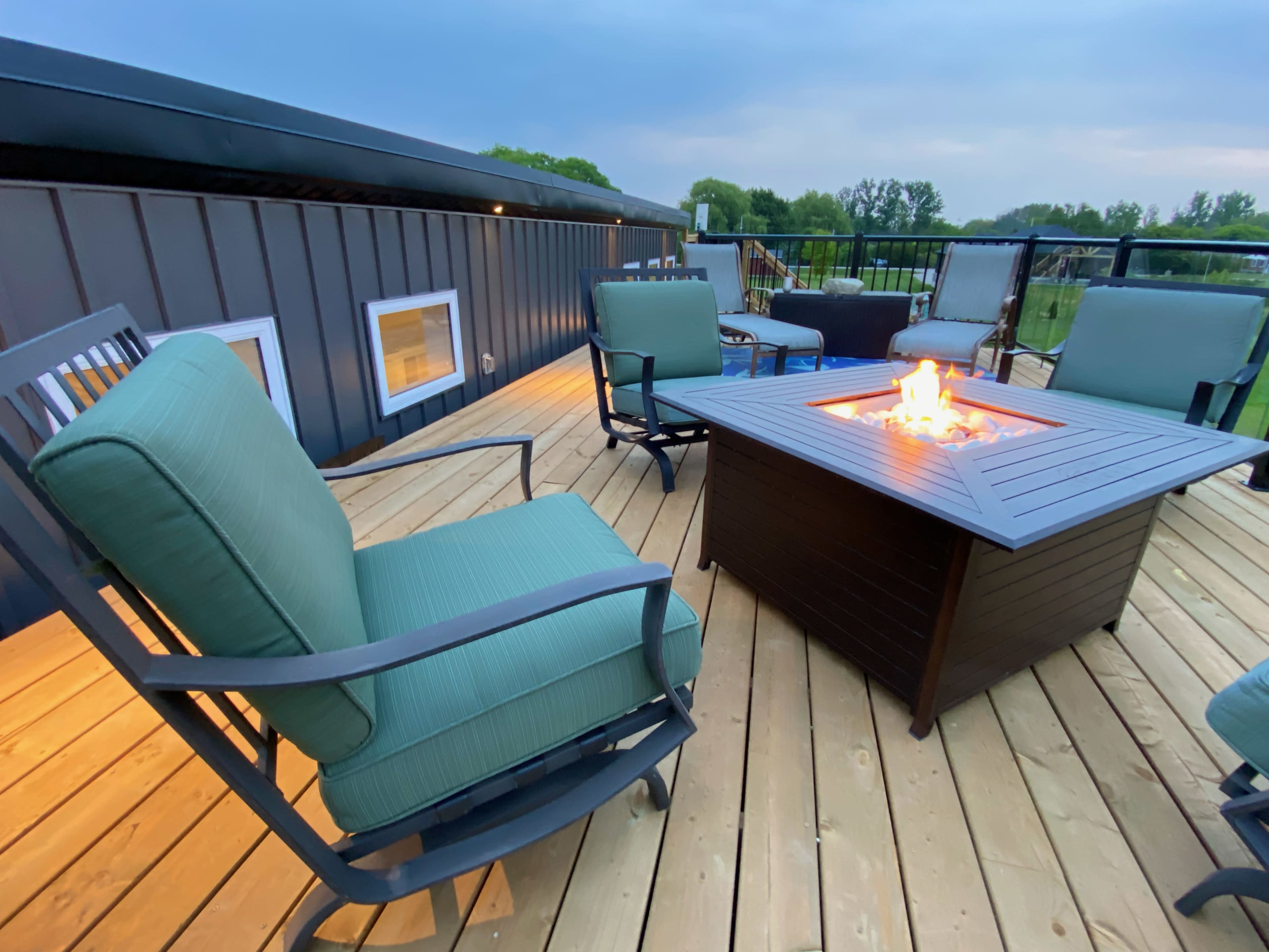 A rectangular fire pit with a flame sits on a wooden deck surrounded by green chairs.