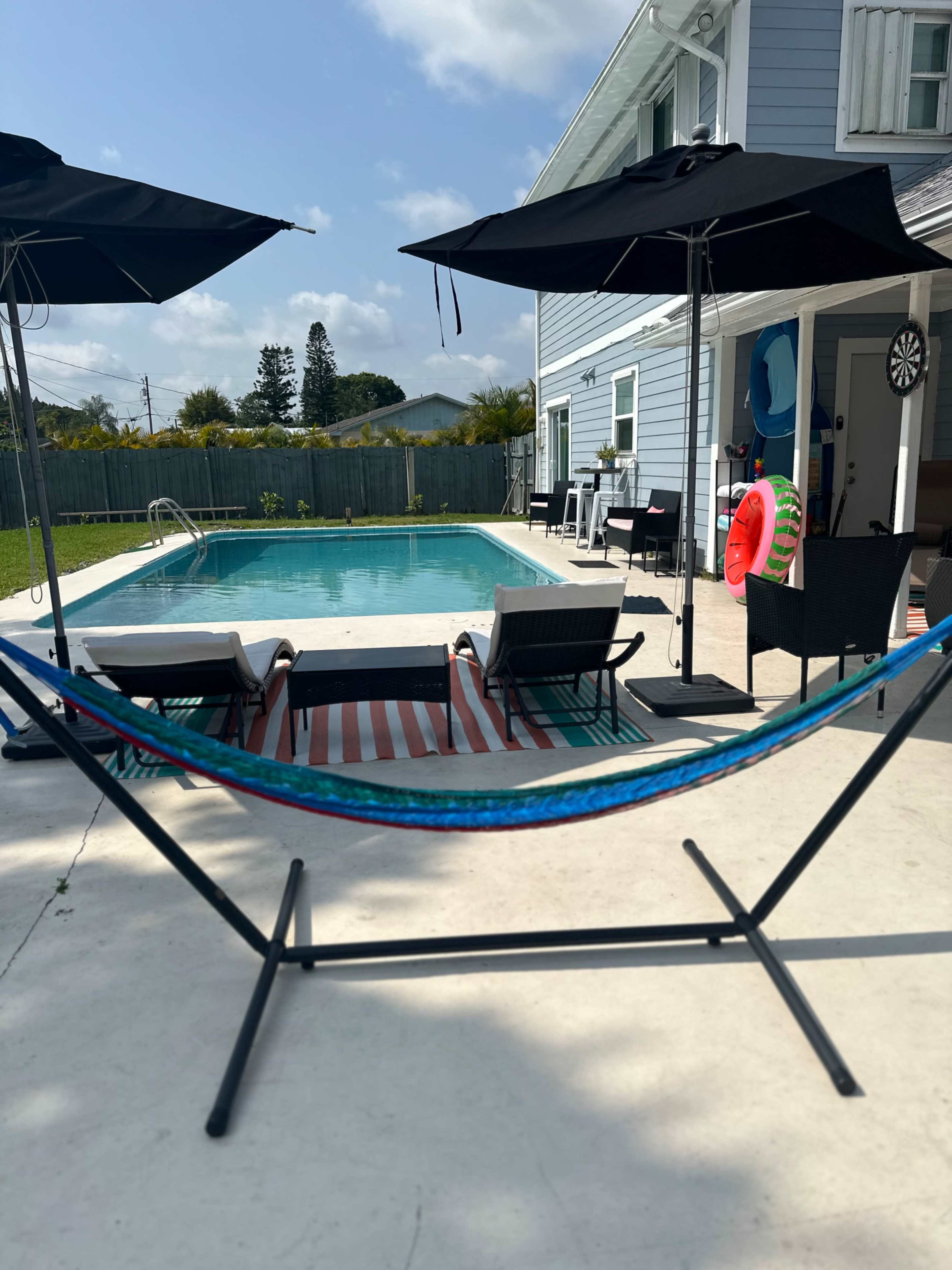 A hammock is positioned in front of a poolside area that features lounge chairs, umbrellas, and a house with blue siding.