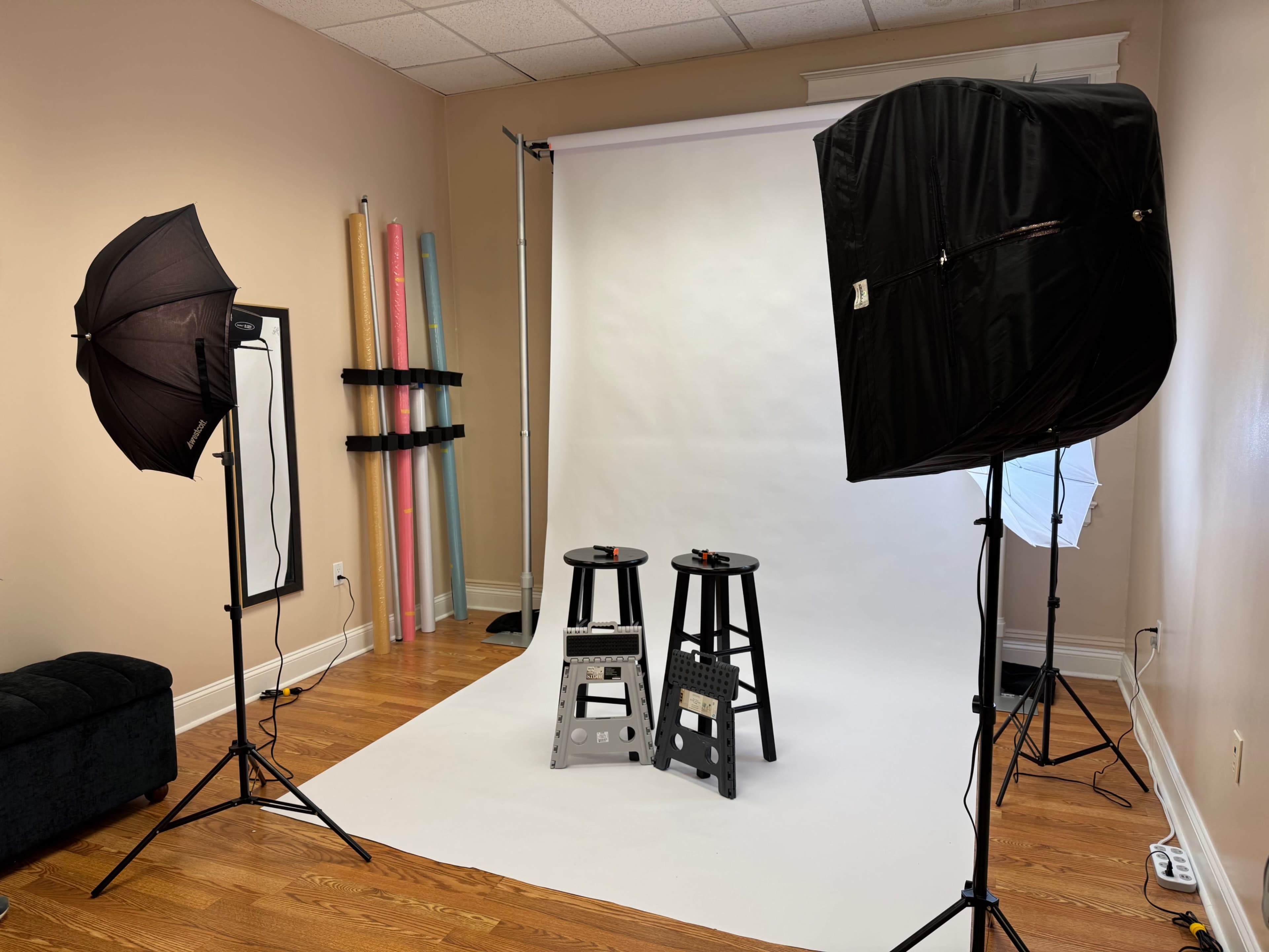 The image shows a photography studio with two black stools positioned on a white backdrop, illuminated by large softbox lights.