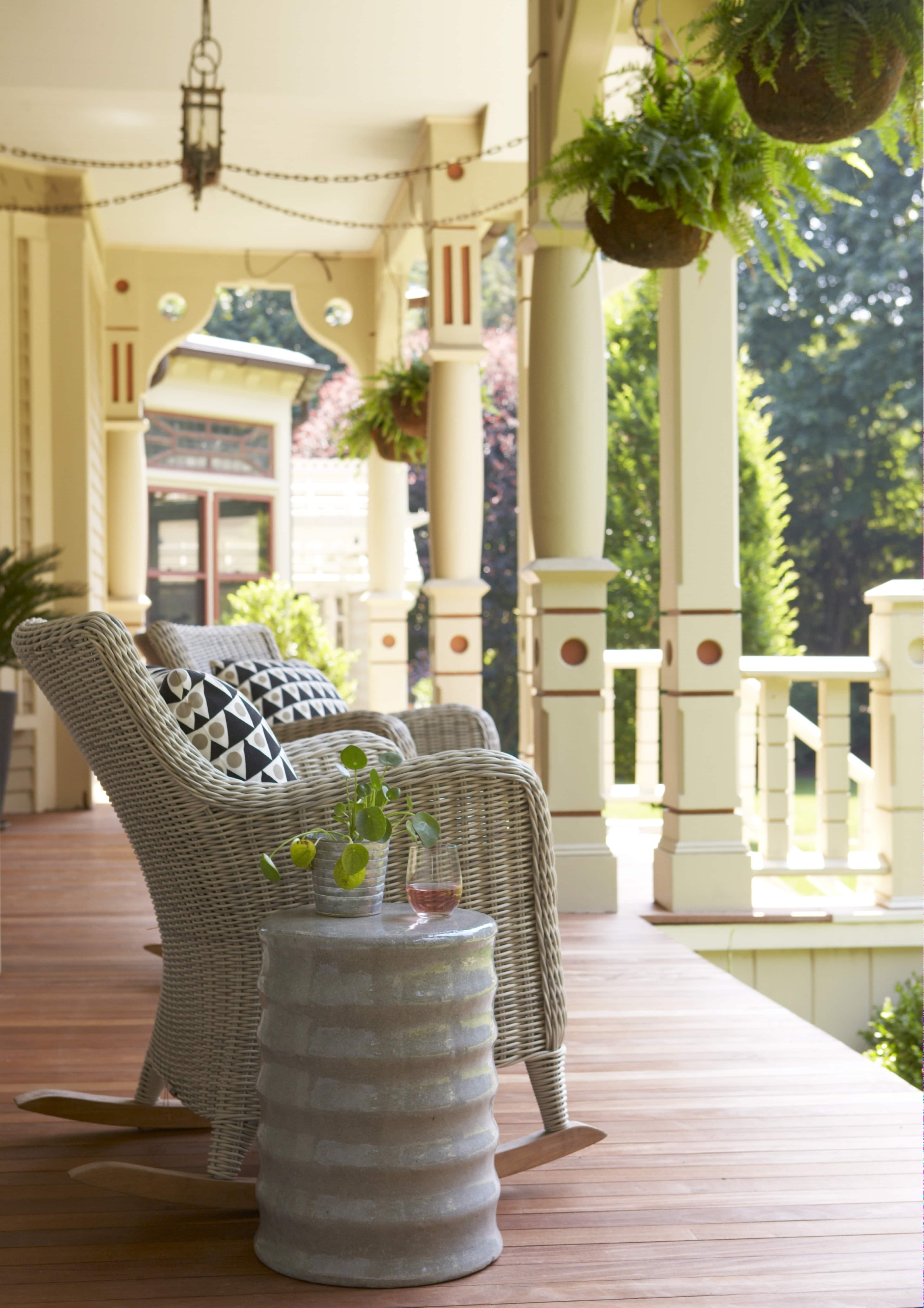 A wicker rocking chair sits on a wooden porch beside a round, textured side table with a small plant and a drink.