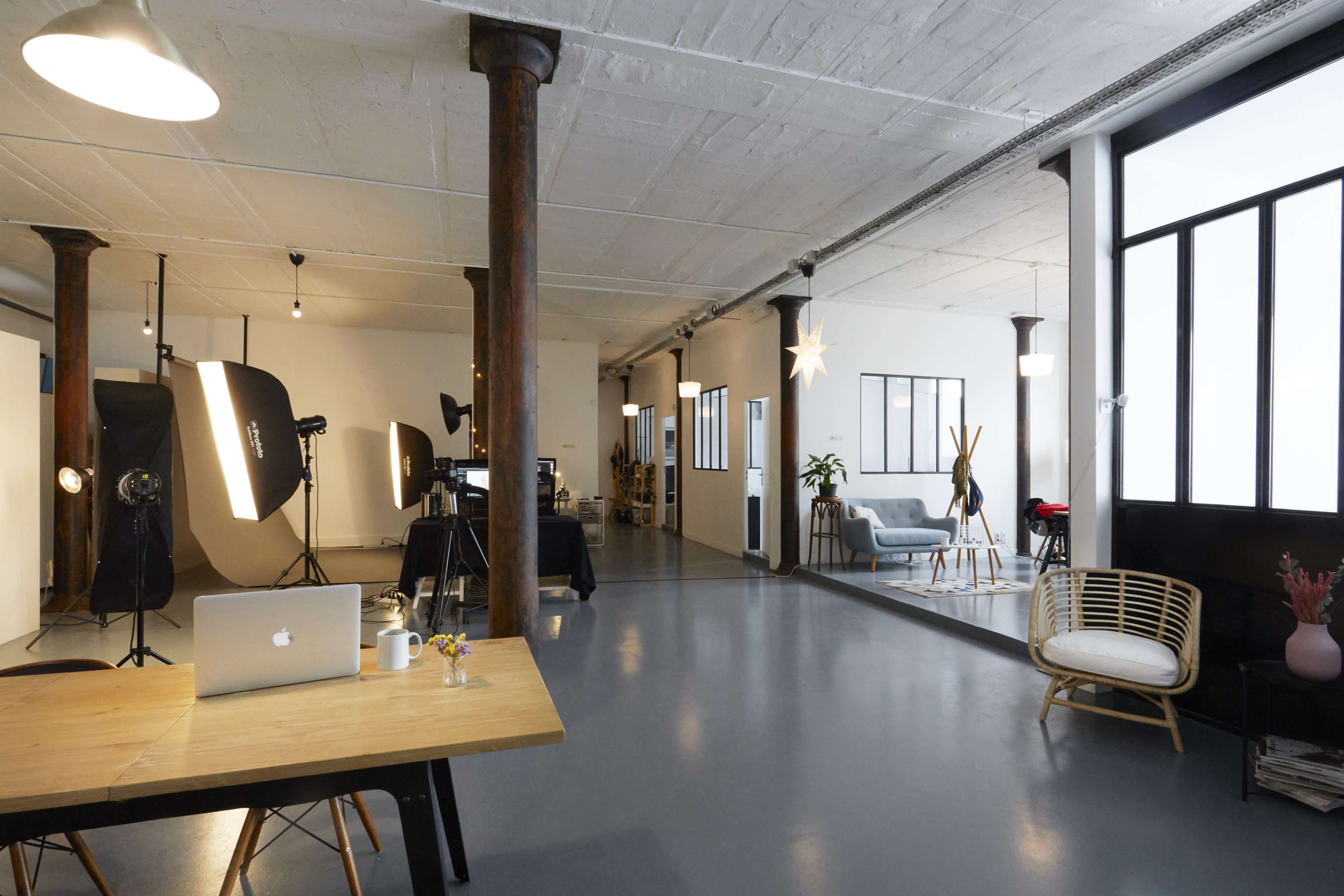 The image shows a spacious, well-lit studio with a modern design, featuring a wooden table with a laptop, seating areas, and photography equipment in the background.