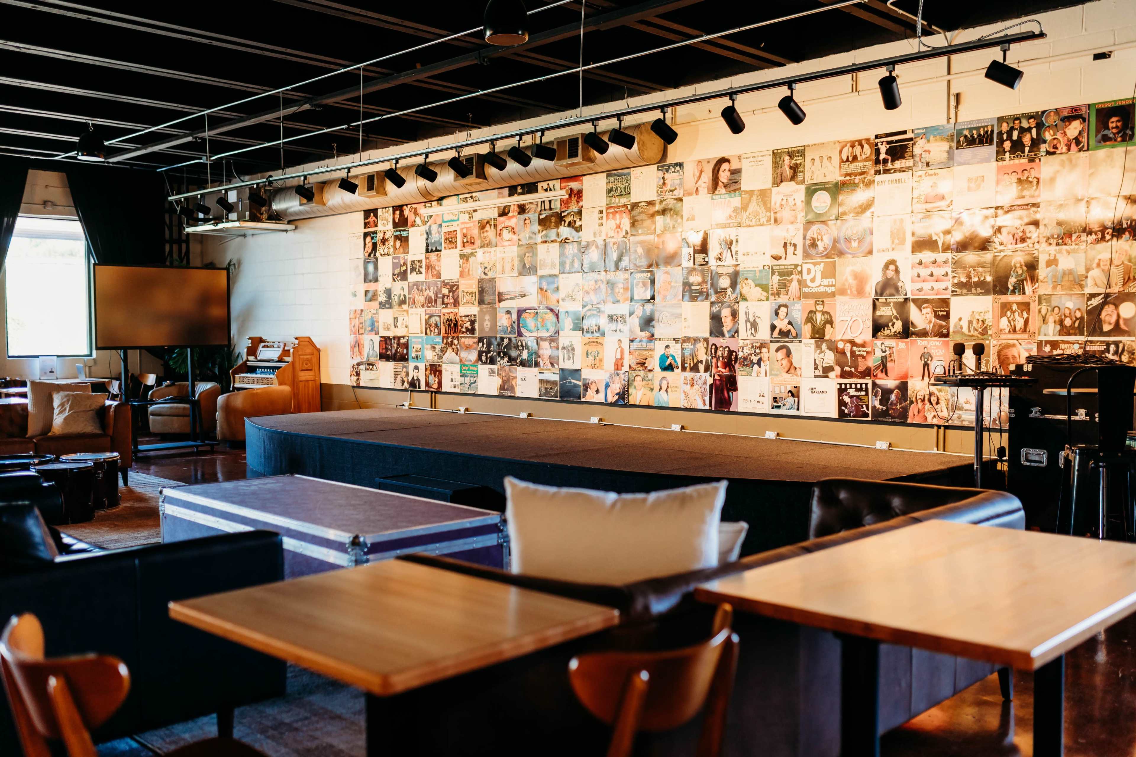 The image shows a stage area in a venue featuring a wall covered with numerous photographs, with seating areas and tables arranged in the foreground.