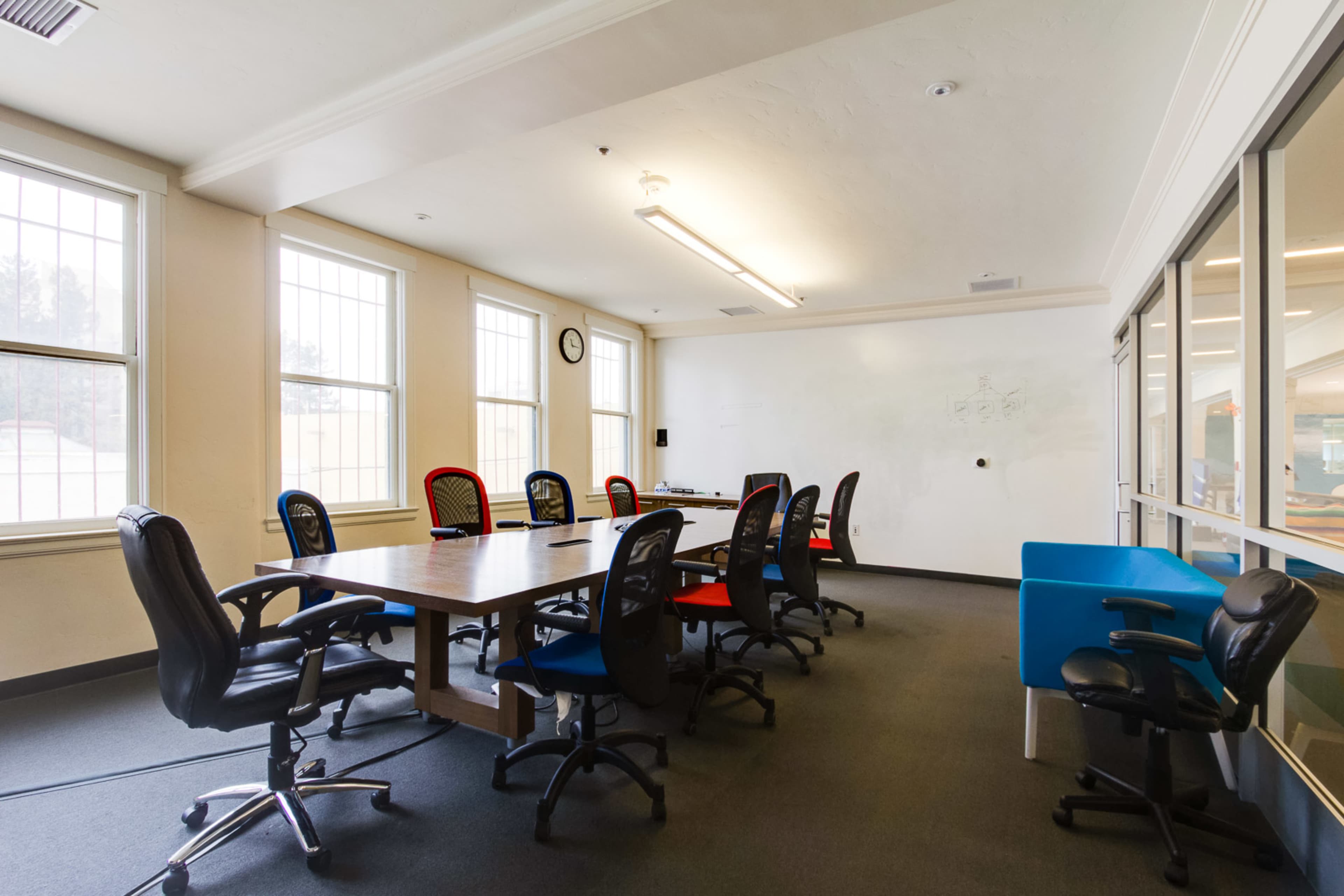 A conference room with a large table surrounded by eight office chairs and two windows letting in light.