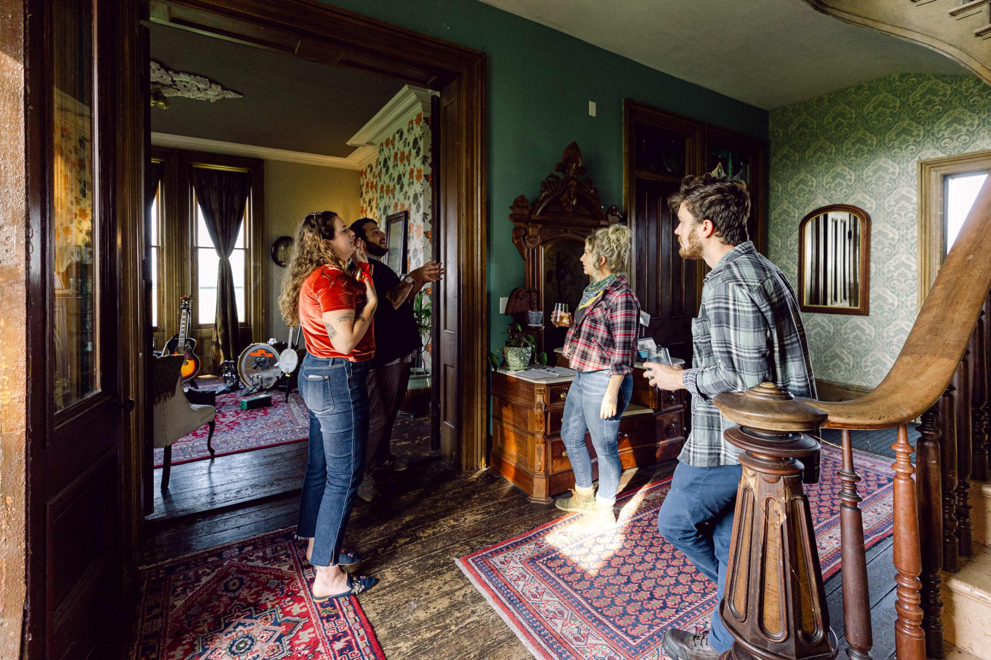 Four people are gathered in a room with vintage decor, chatting and enjoying drinks near an ornate wooden staircase.