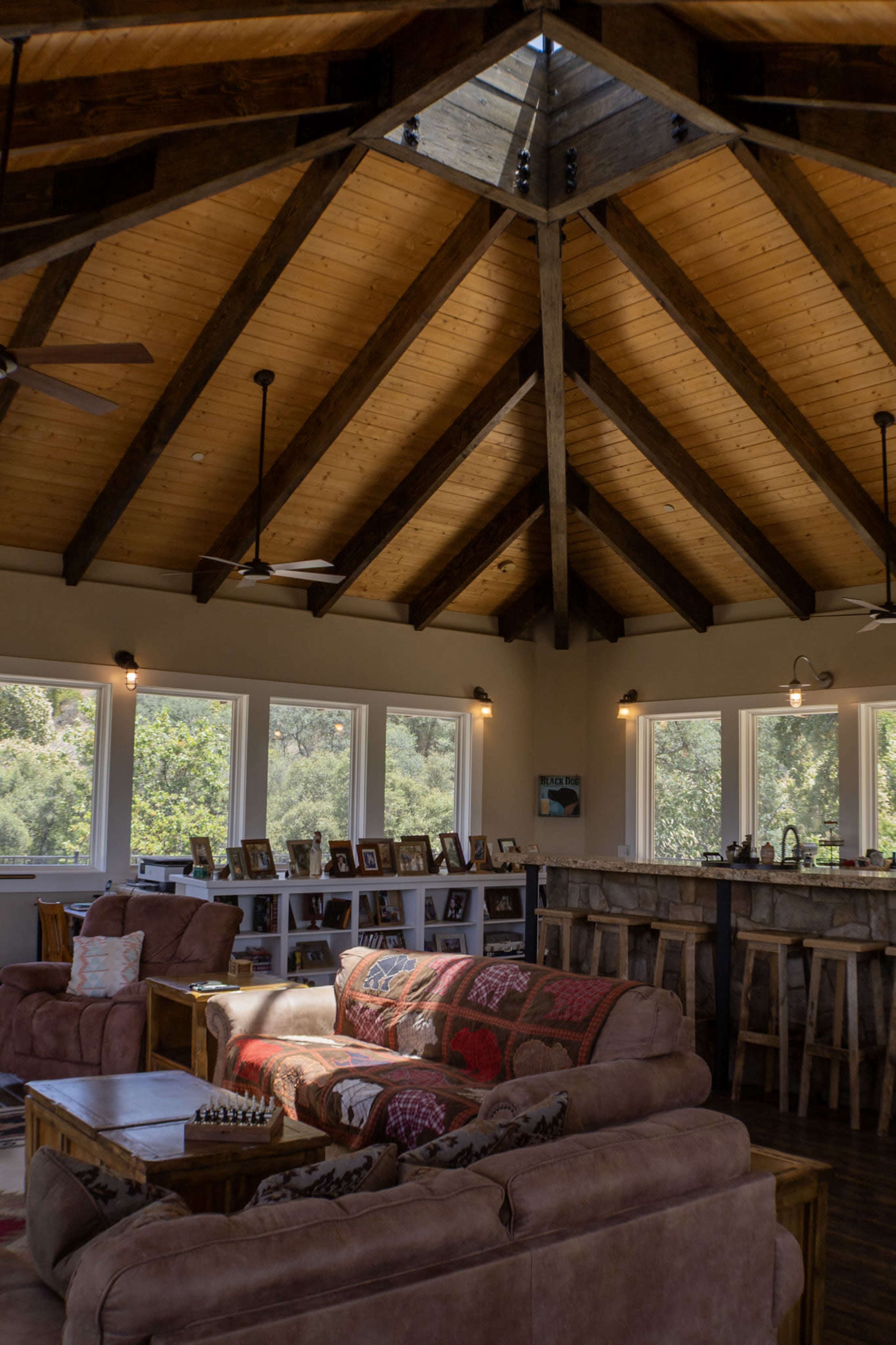 The image shows a spacious living area with a high, wooden-beamed ceiling, featuring comfortable seating and a bar area with several bar stools.
