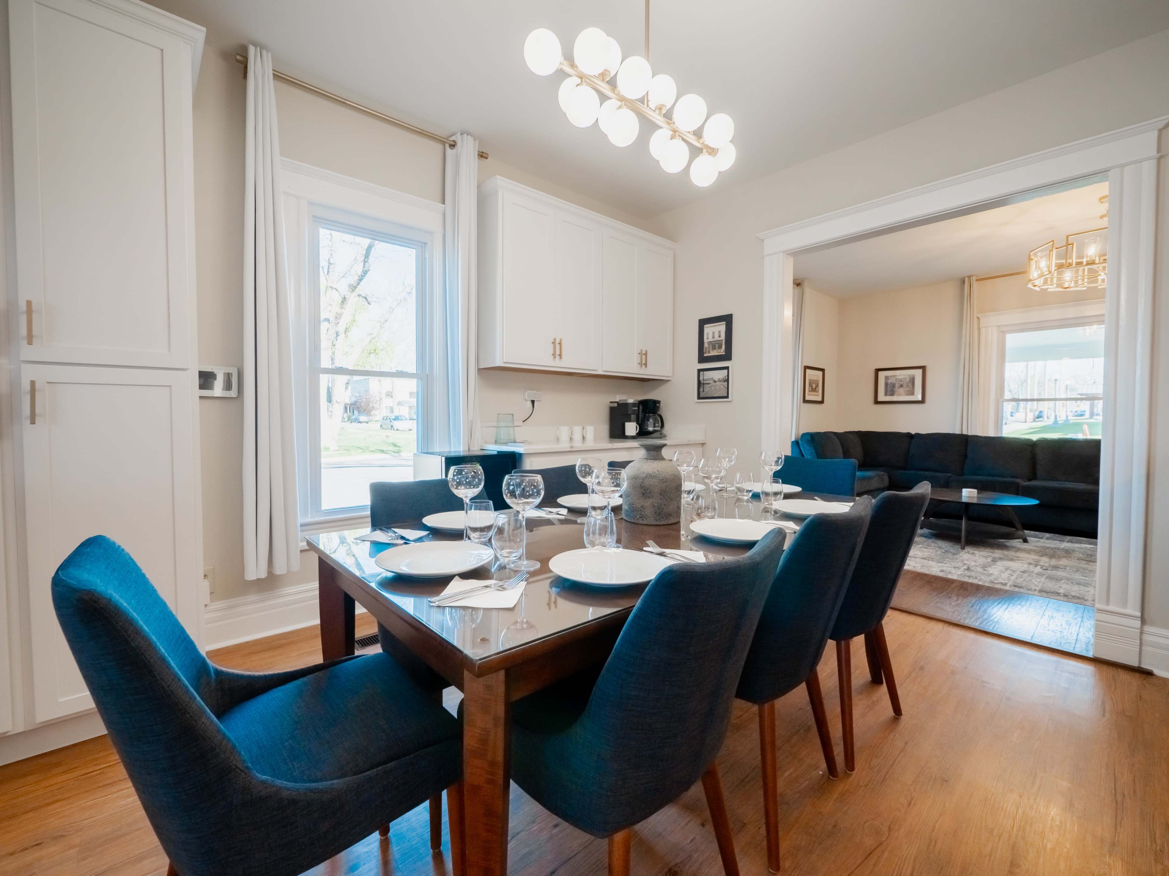 A modern dining room features a wooden table with blue upholstered chairs, set for a meal, and a bright window overlooking the outside.