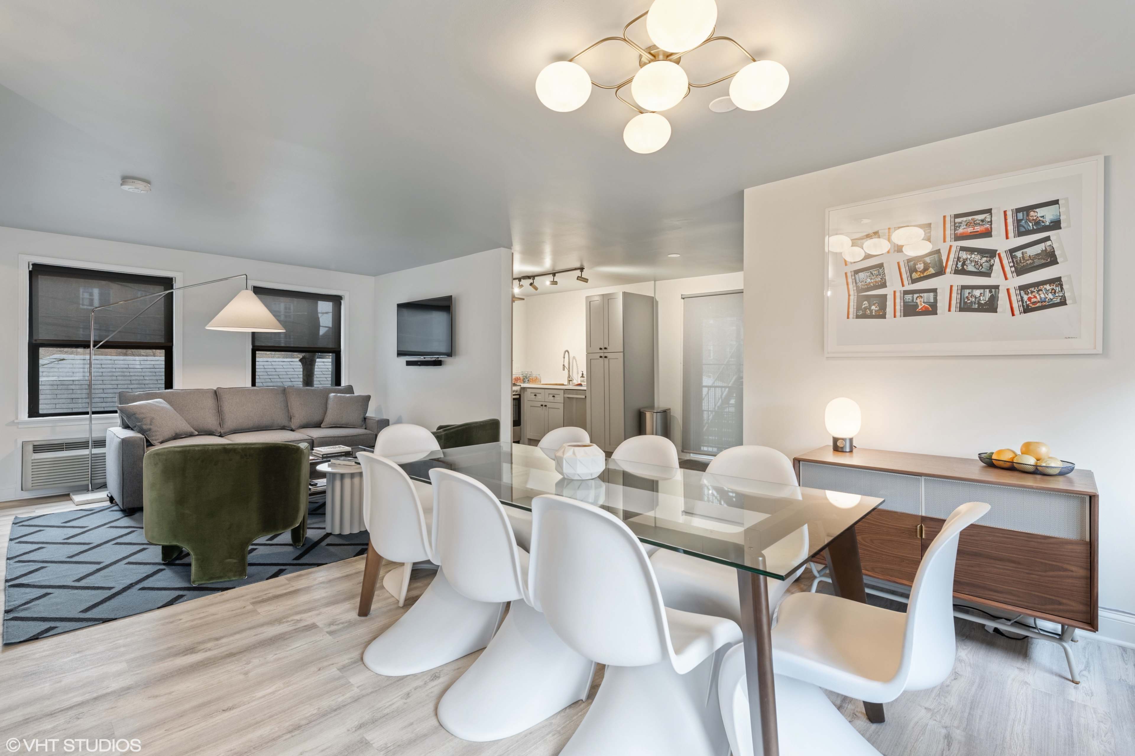 A bright, modern living and dining area featuring a glass dining table with white chairs, a sofa, and a kitchenette.