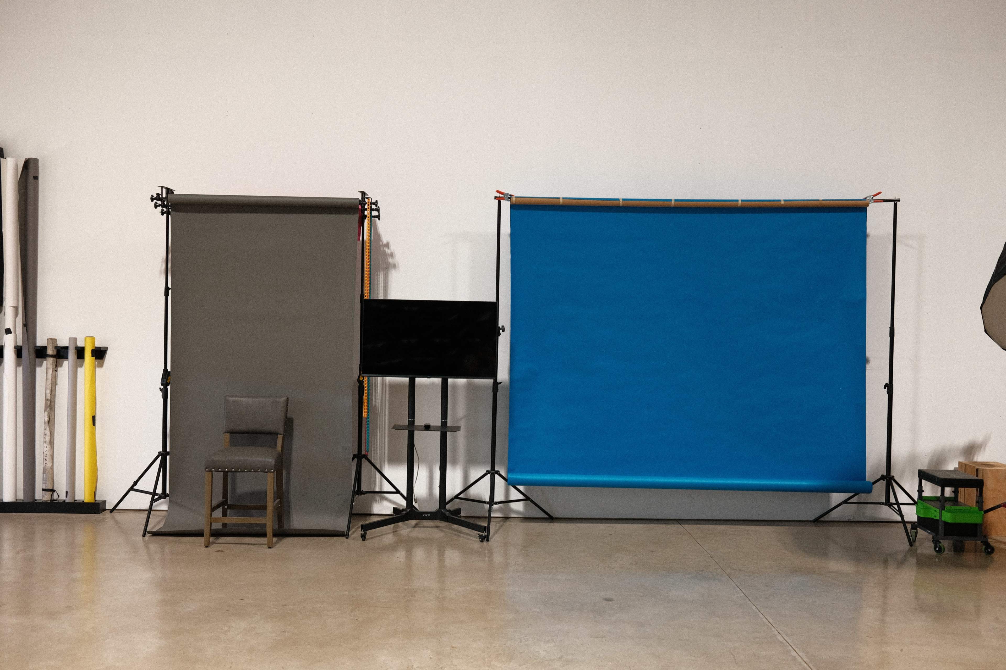 A photography studio setup with a gray background, a blue backdrop, a chair, and a monitor on stands.