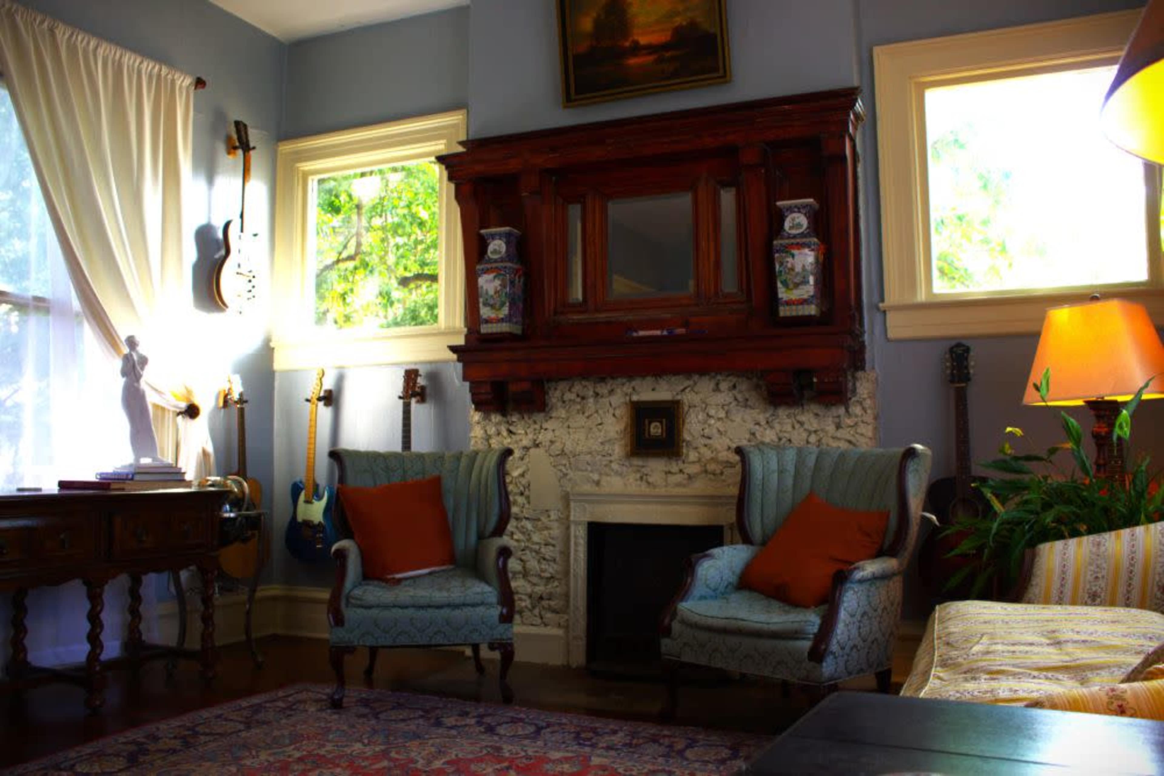 The image shows a cozy living room with two patterned armchairs, a wooden fireplace surrounded by stone, a small table, and various guitars displayed on the walls.