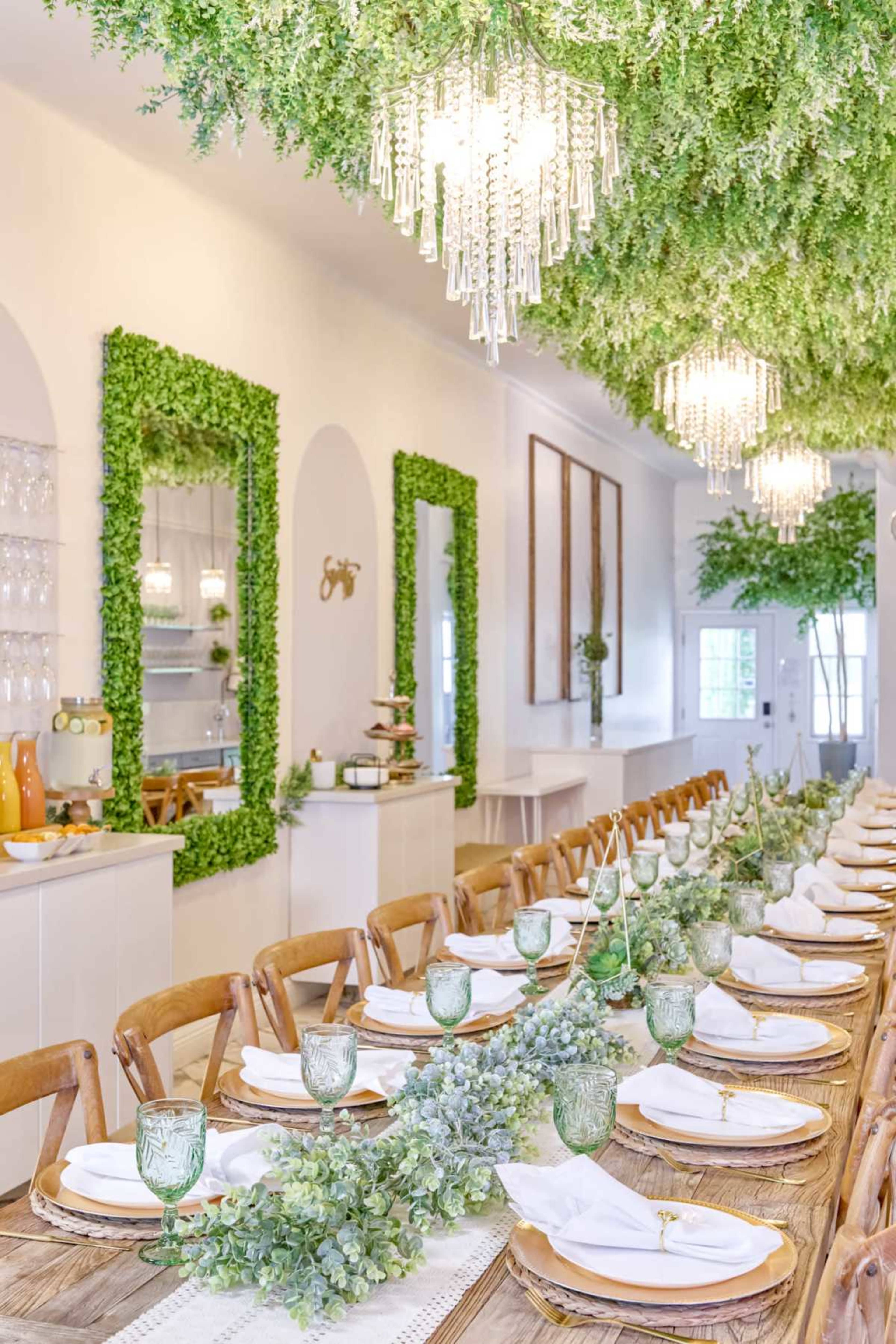 A long dining table is set with white plates and green glassware, surrounded by walls adorned with greenery and chandeliers hanging above.
