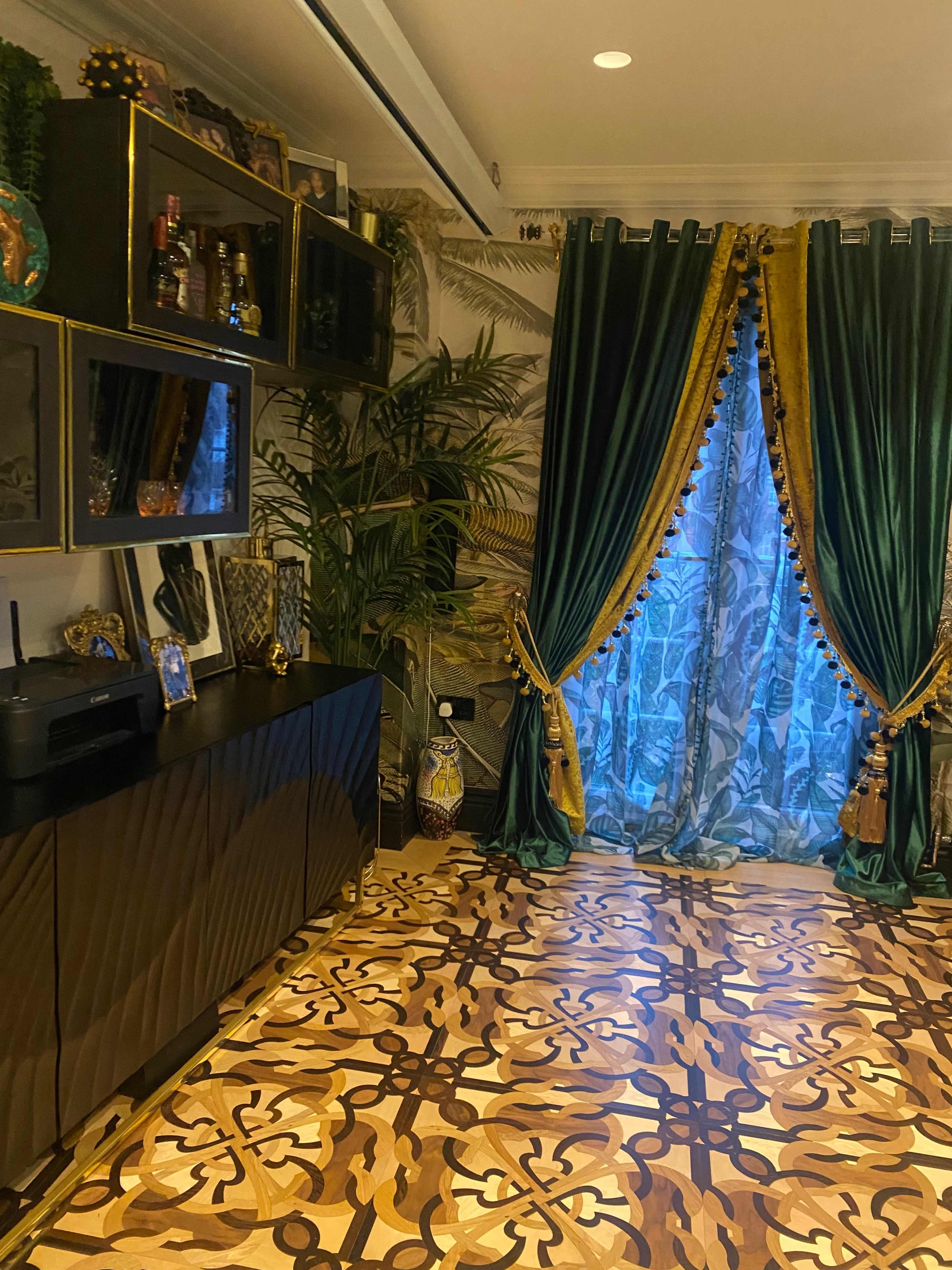 A room with a patterned wooden floor, dark cabinetry, and elegant green drapes adorned with gold trim, framed by tropical-themed wallpaper.