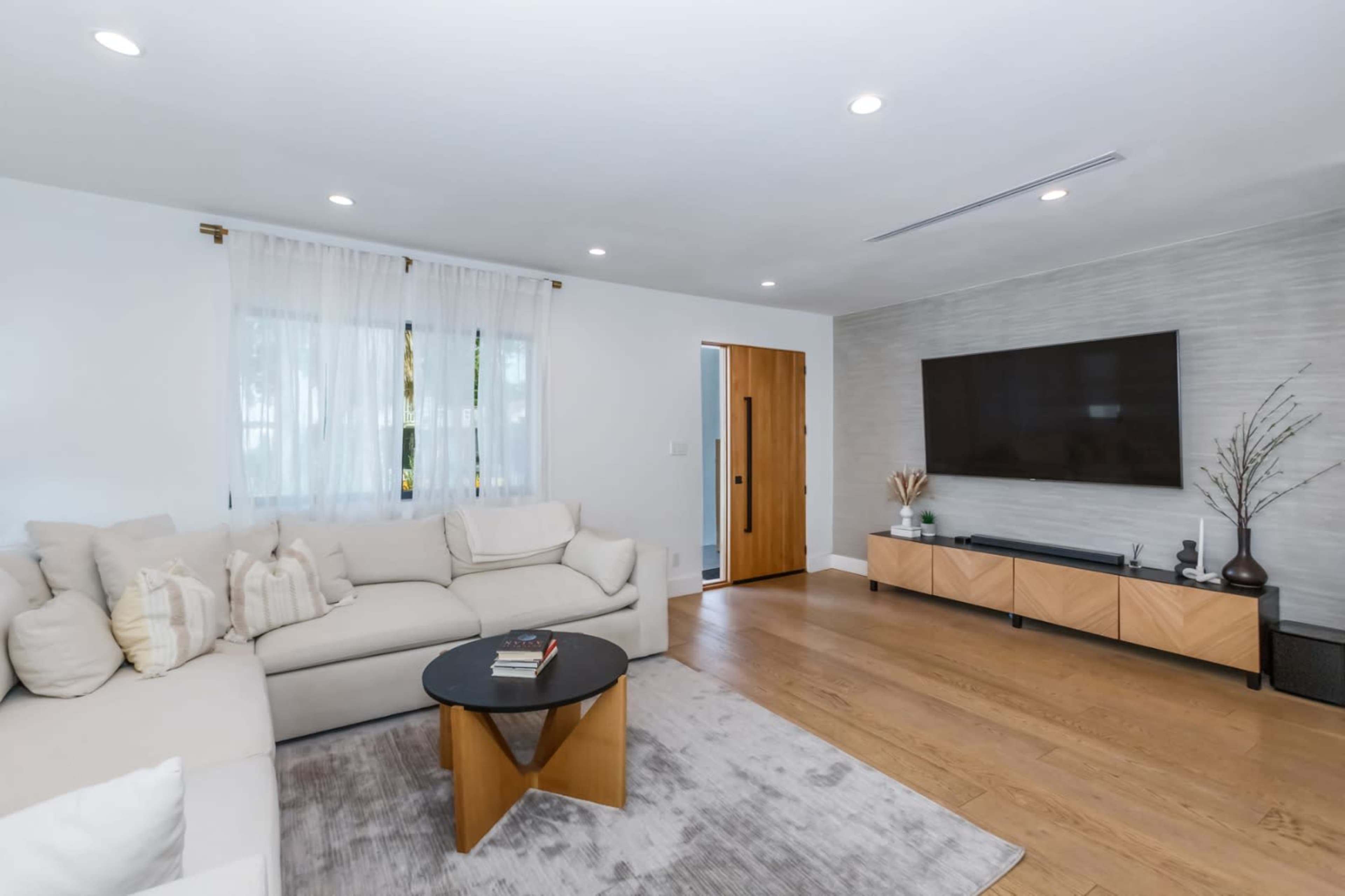 A modern living room featuring a sectional couch, a round coffee table, wooden flooring, a wall-mounted TV, and minimal decor.