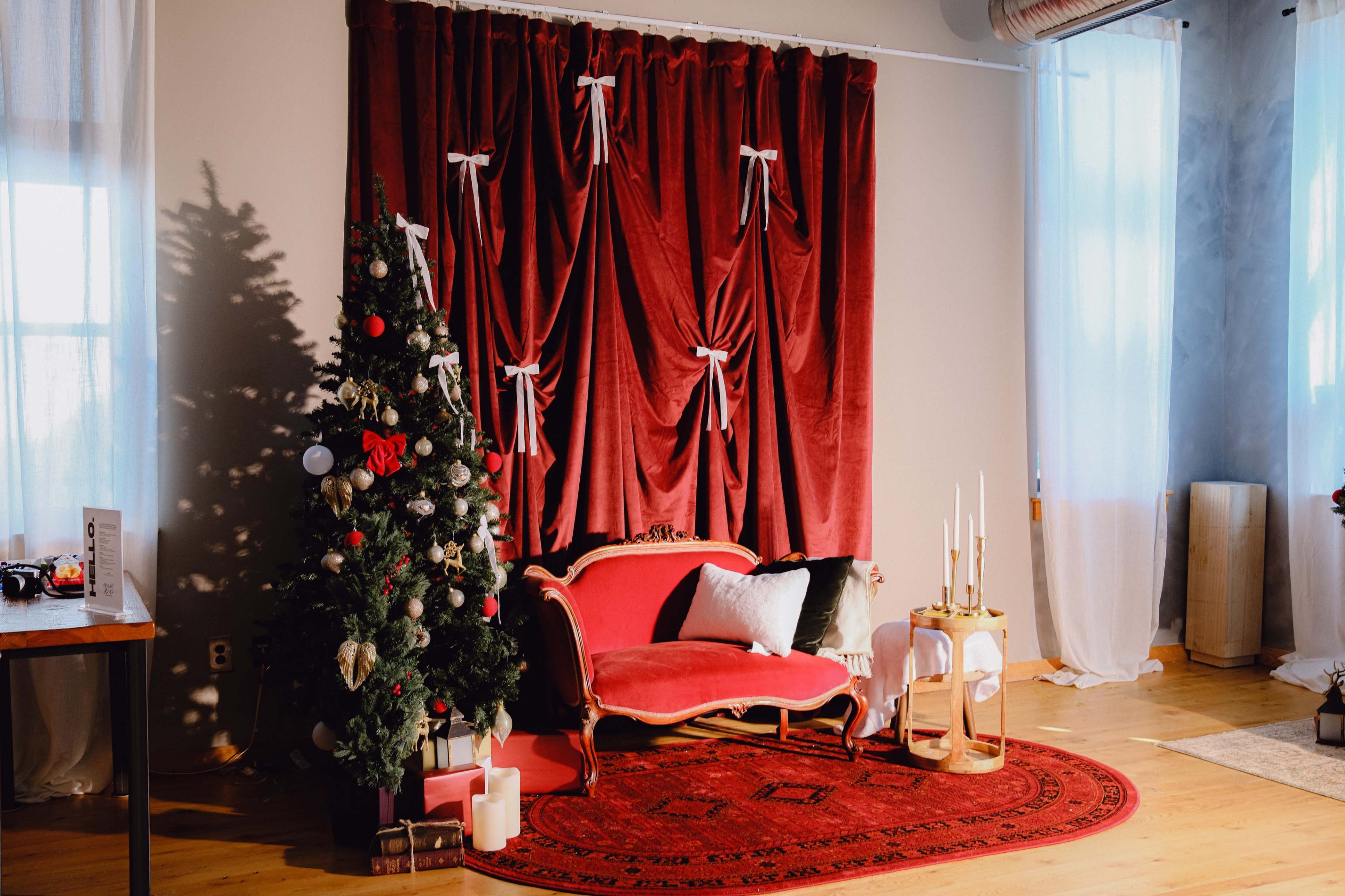 A red velvet couch is displayed in front of a backdrop of draped red fabric, accompanied by a Christmas tree decorated with ornaments and candles nearby.