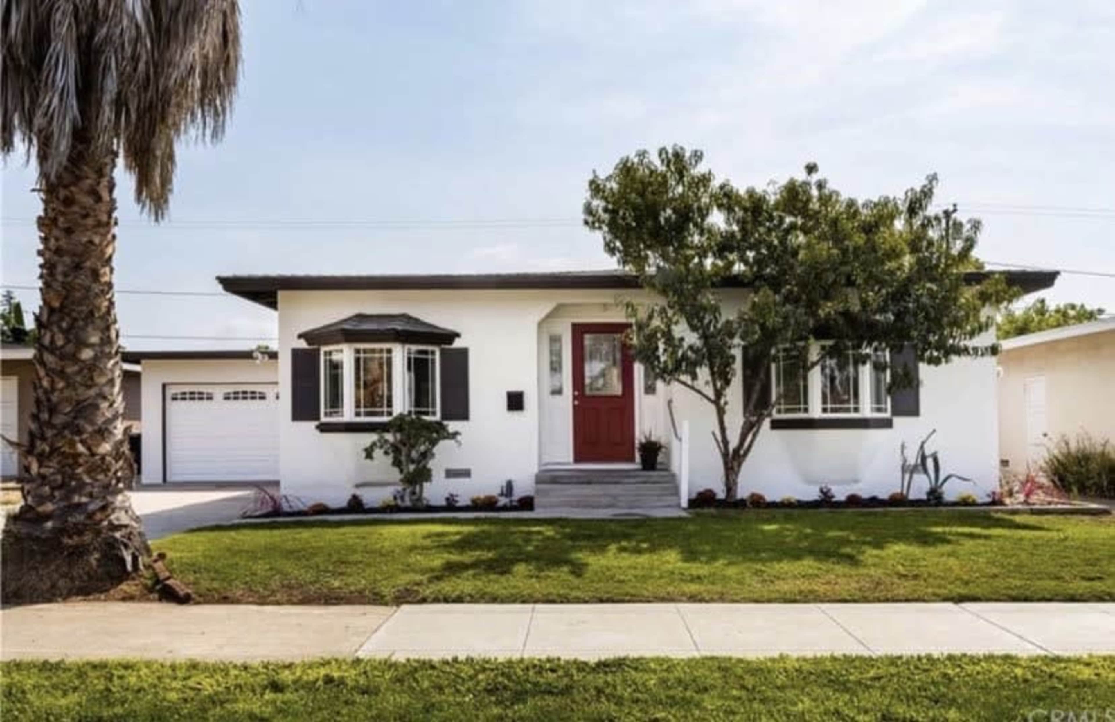 The image shows a single-story, white house with a red door, surrounded by green grass and a palm tree in the front yard.