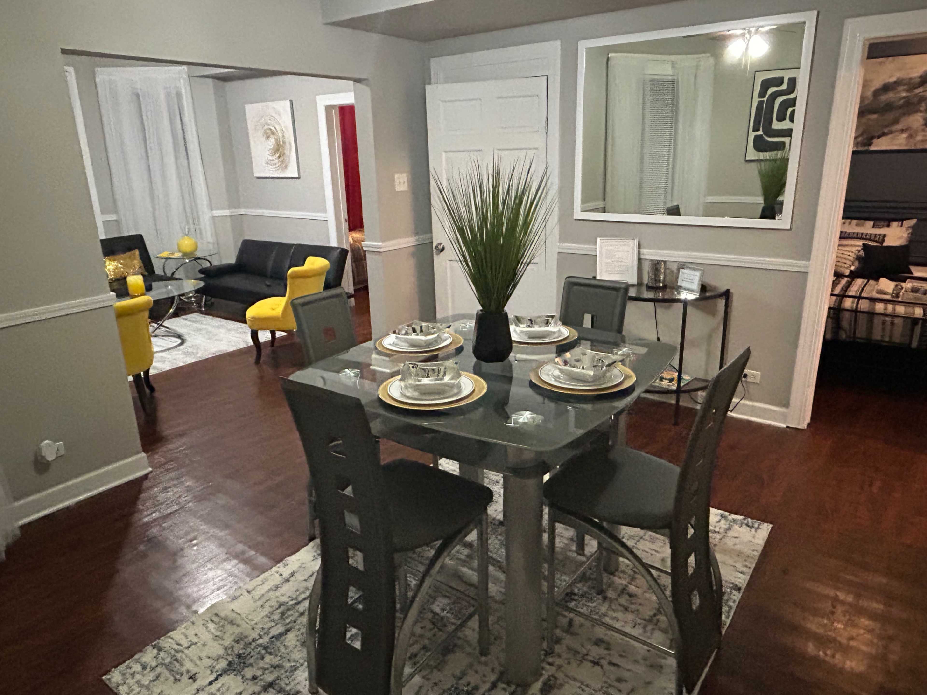 The image shows a modern dining area with a glass table set for four, surrounded by gray chairs, adjacent to a living space featuring a black couch and yellow chairs.