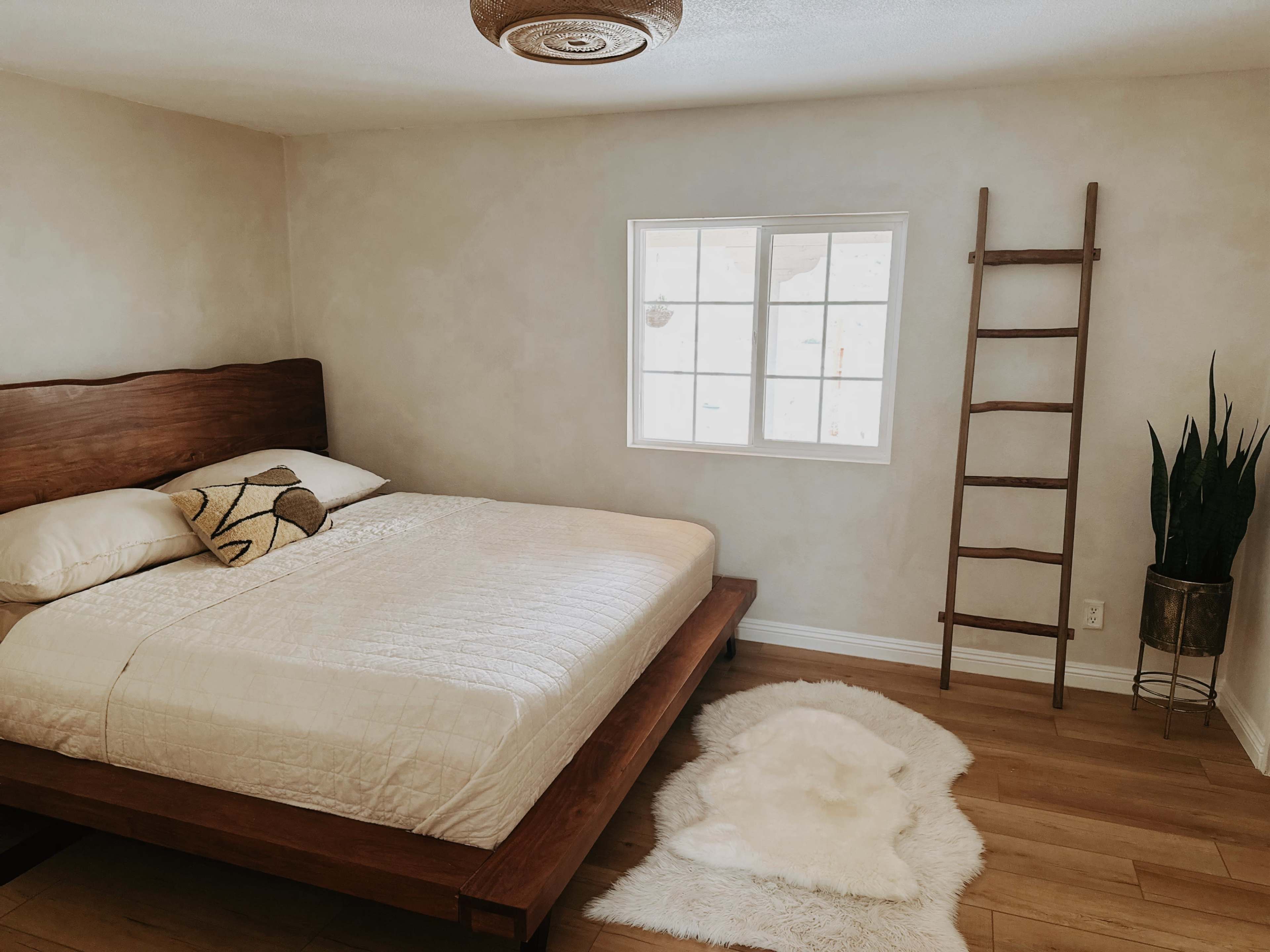 A simple bedroom featuring a wooden bed, a window, a decorative ladder, and a potted plant.