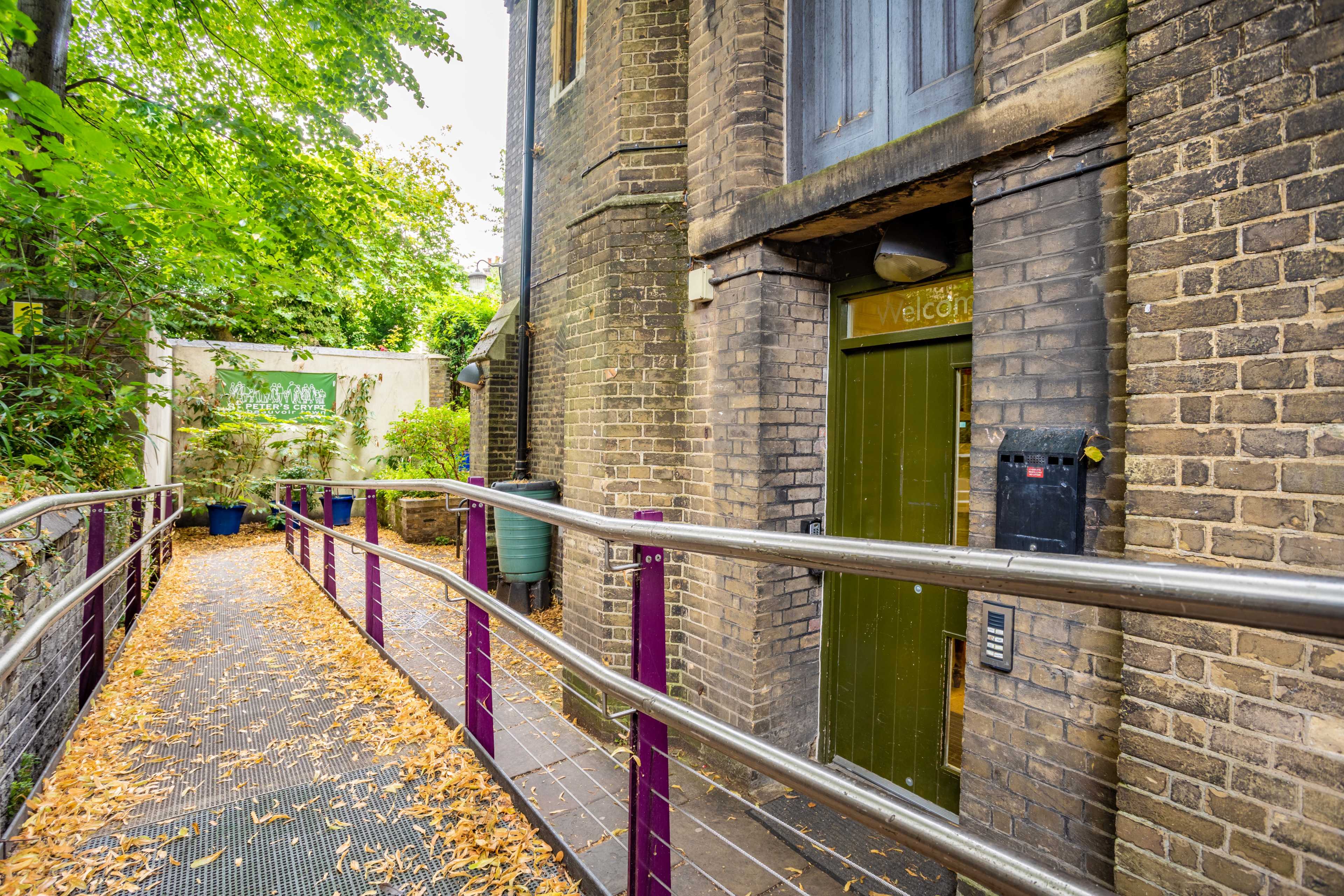 A narrow pathway lined with fallen leaves leads to a green door marked "welcome" at the entrance of a brick building.