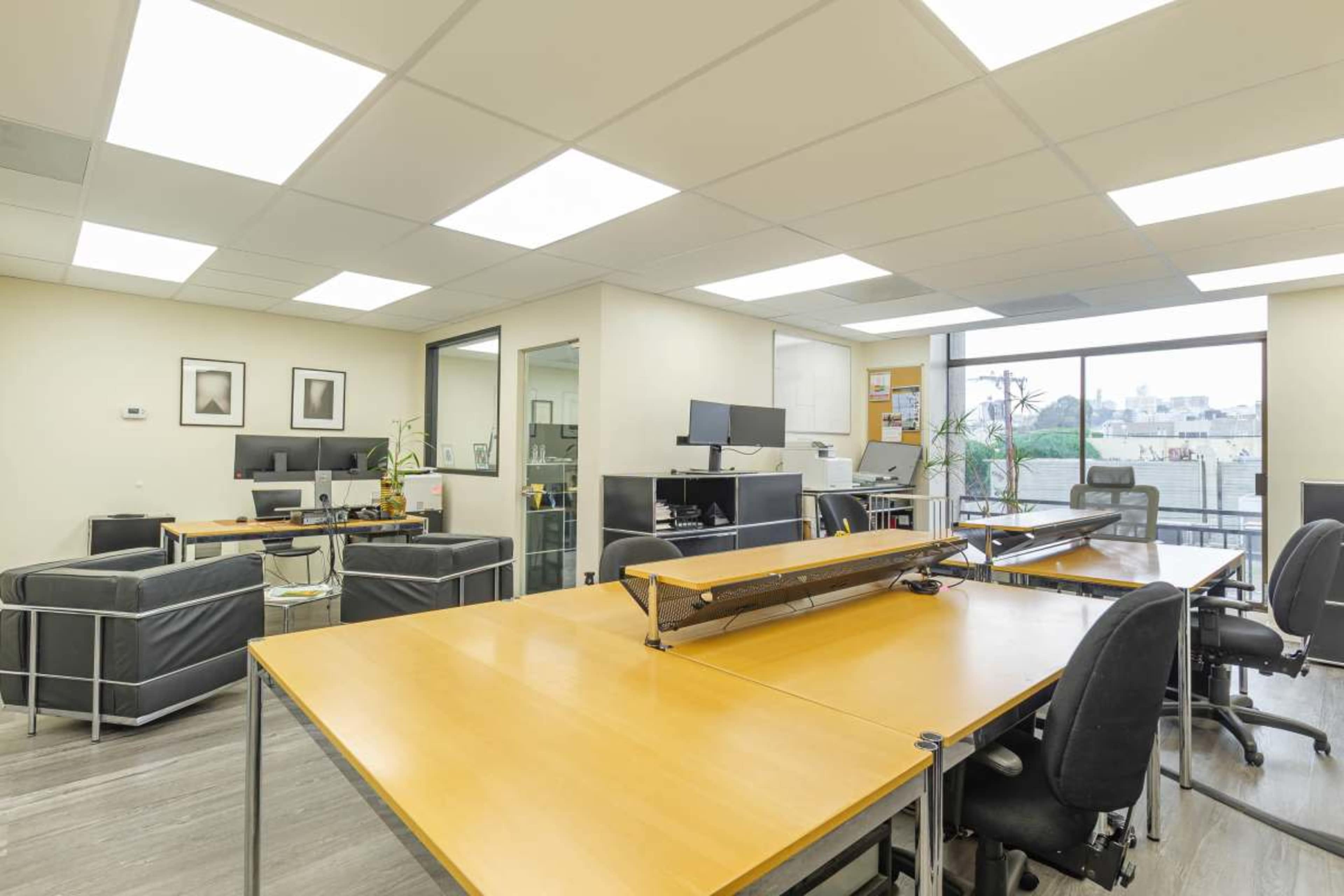 The image shows a modern office space with multiple desks, computer stations, and large windows allowing natural light.